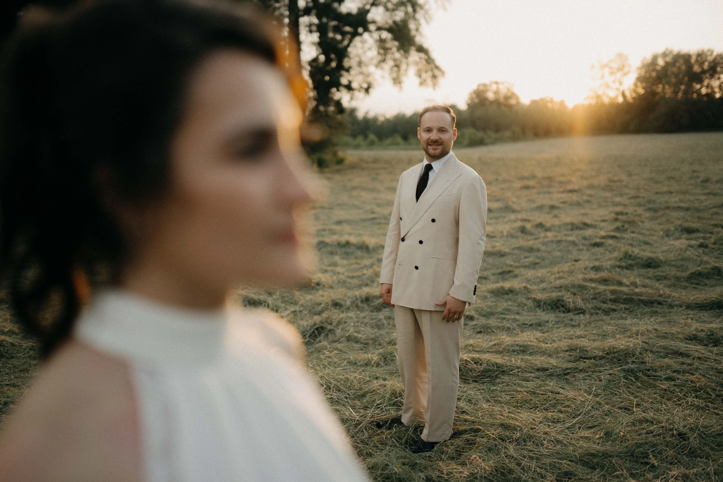 A man in a beige suit and black tie smiling at the camera in an outdoor field during sunset, with a blurred woman in the foreground.