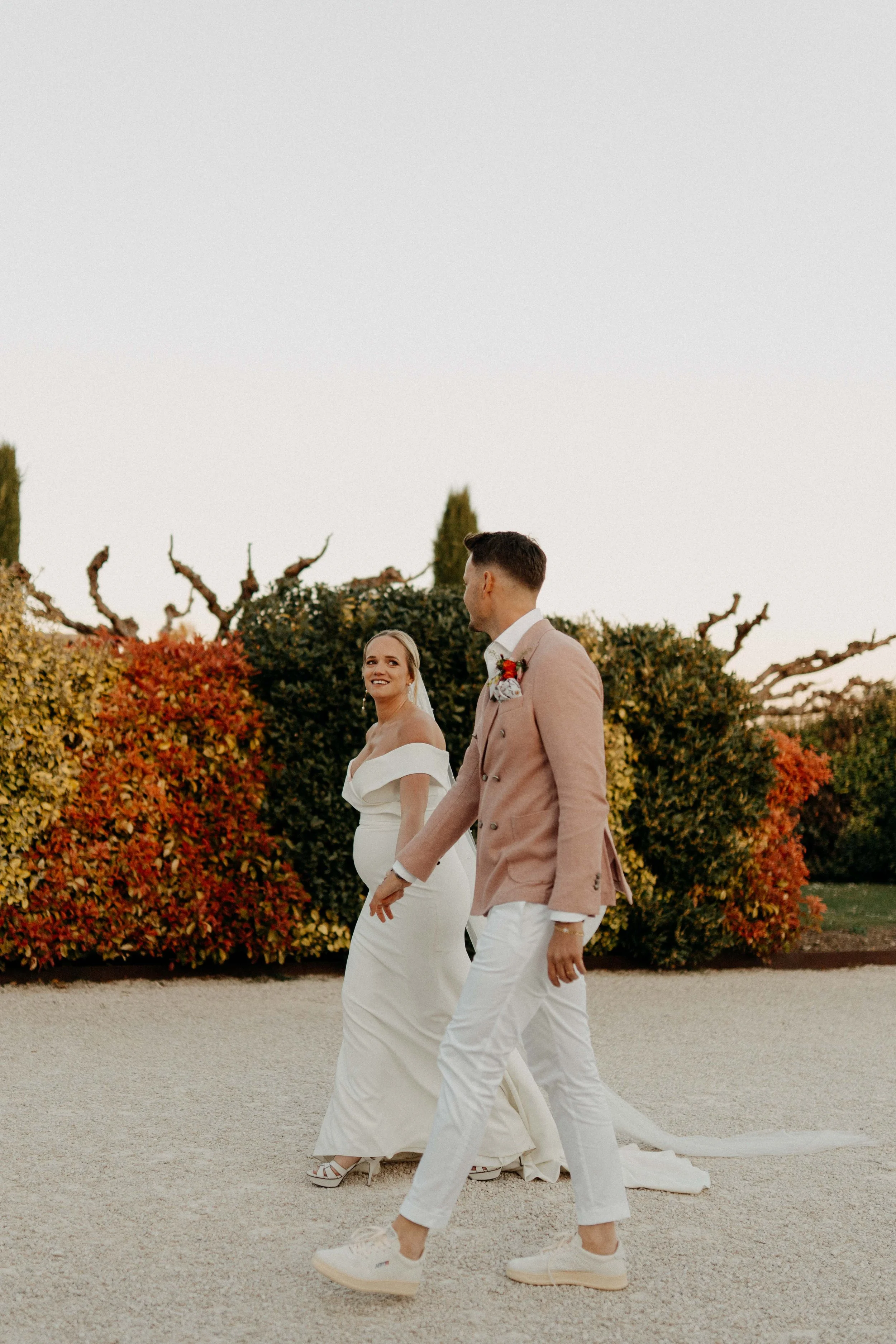 A bride and groom holding hands and walking in a garden with autumn-colored bushes in the background during sunset.