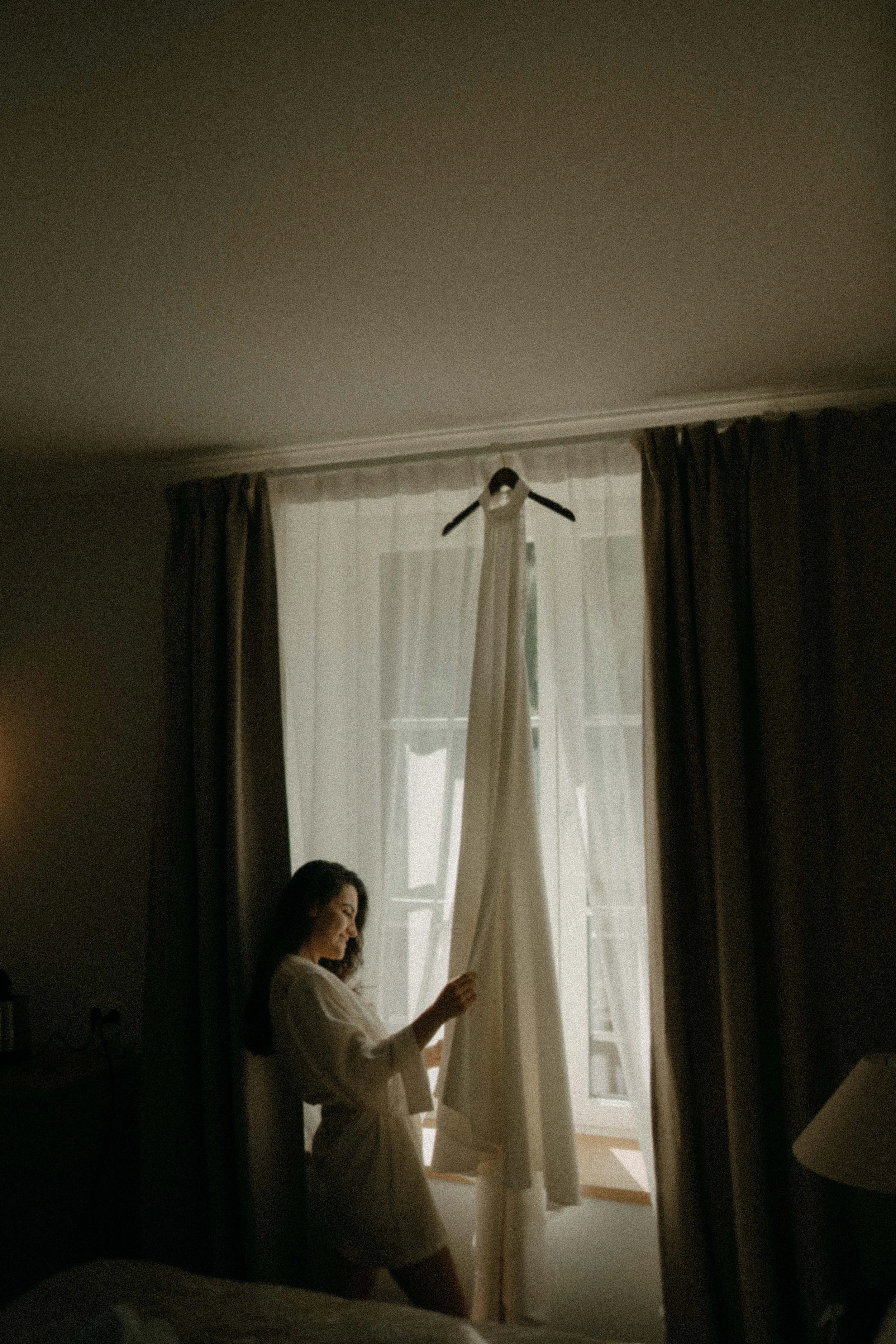 A woman in a white shirt and beige shorts standing by a window with curtains, looking at her phone. Sunlight filters through the sheer curtains.