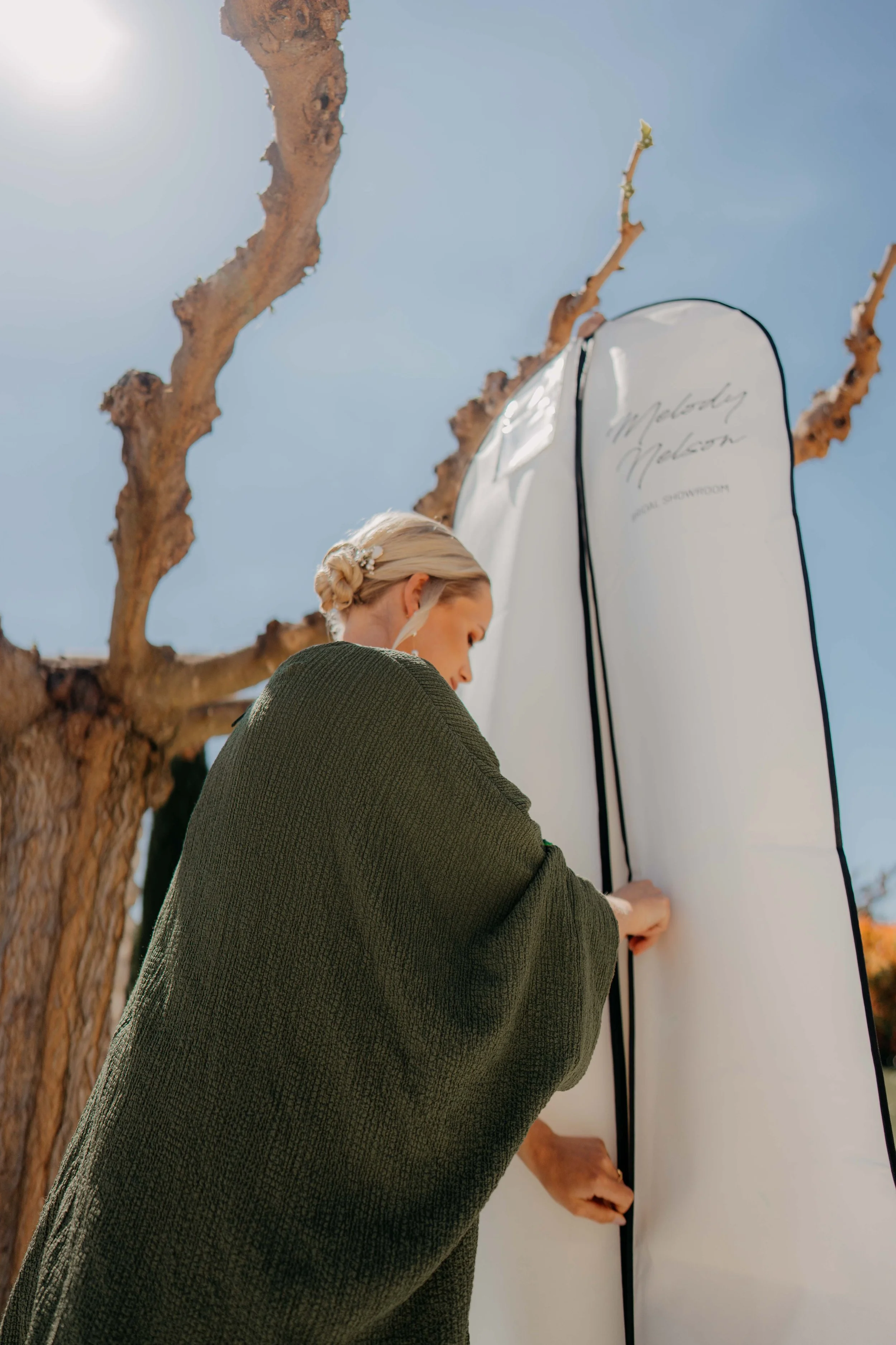 A woman with blonde hair styled in a low bun, wearing a green textured jacket, standing outside under a leafless tree with a clear blue sky background. She is holding a large white card or board with the words 'Melody Melson' and 'Flower Download' wr