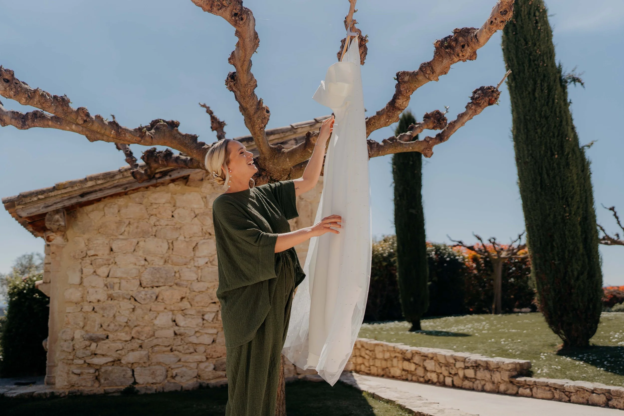 A smiling woman with blonde hair, wearing a dark green dress, is hanging white fabric or curtain on a tree branch outside near a stone building and tall cypress trees under a clear blue sky.