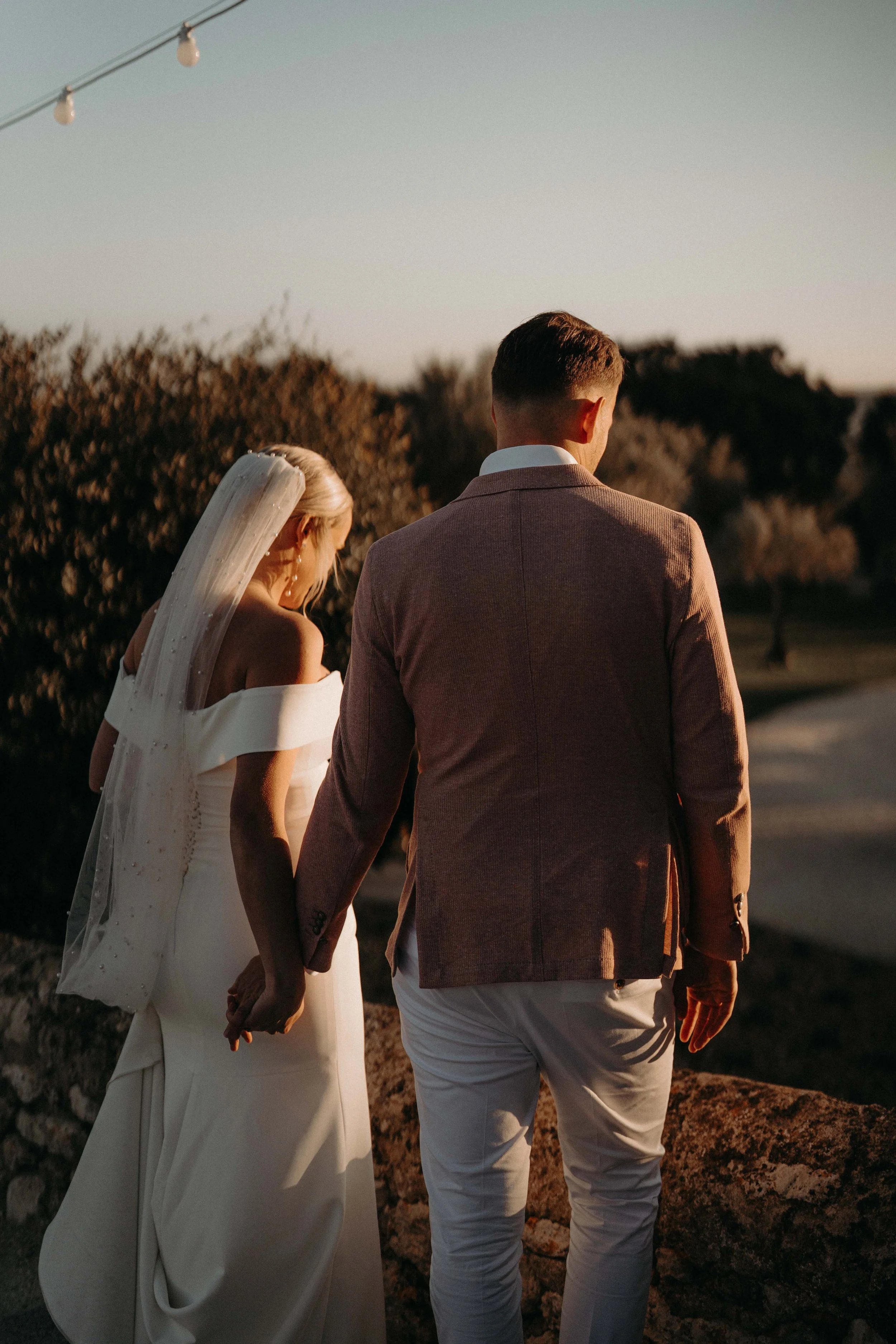 A bride and groom holding hands, walking outdoors during sunset, with the bride in a white dress and veil and the groom in a brown blazer and light-colored pants.