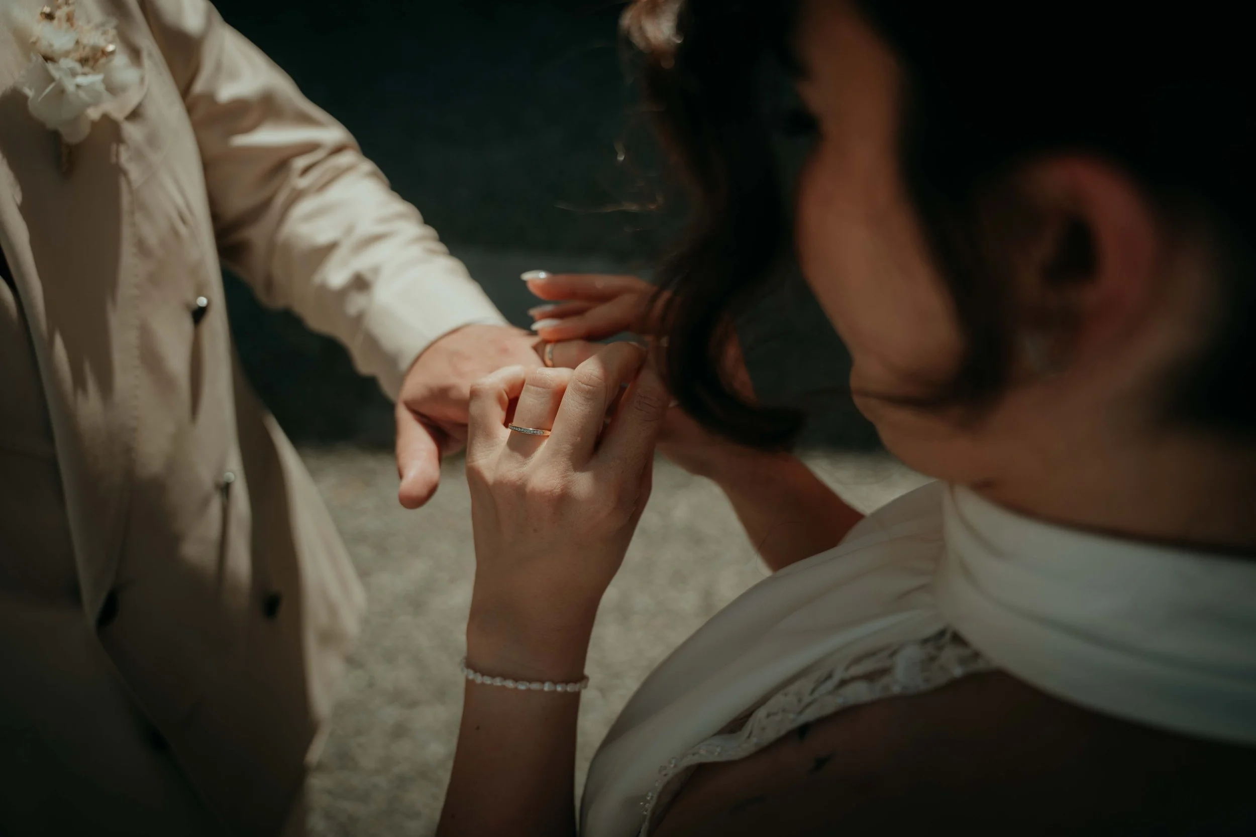 A woman with dark hair and a pearl bracelet kneels down to kiss the hand of another person, who is wearing a cream-colored outfit with decorative flowers and a ring.