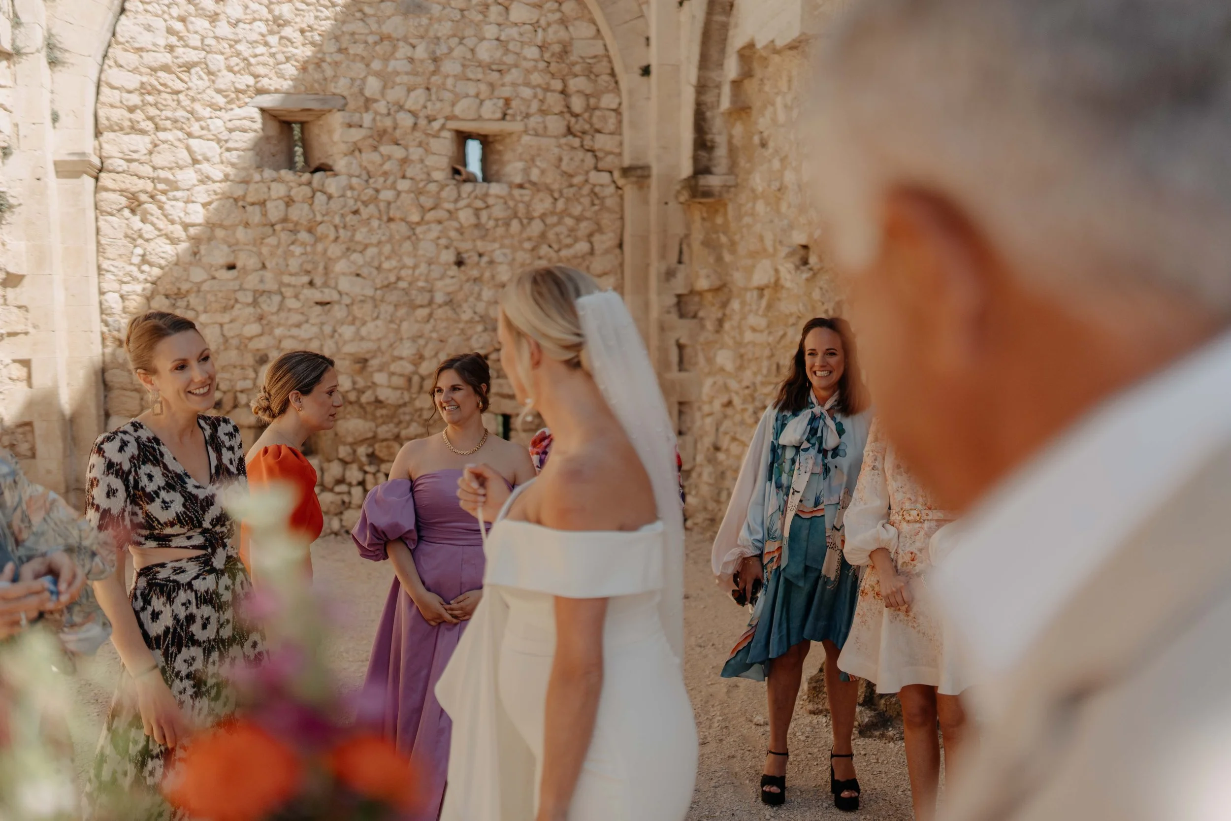 A group of women in colorful dresses smiling and talking at an outdoor gathering in front of a rustic stone wall with small windows.