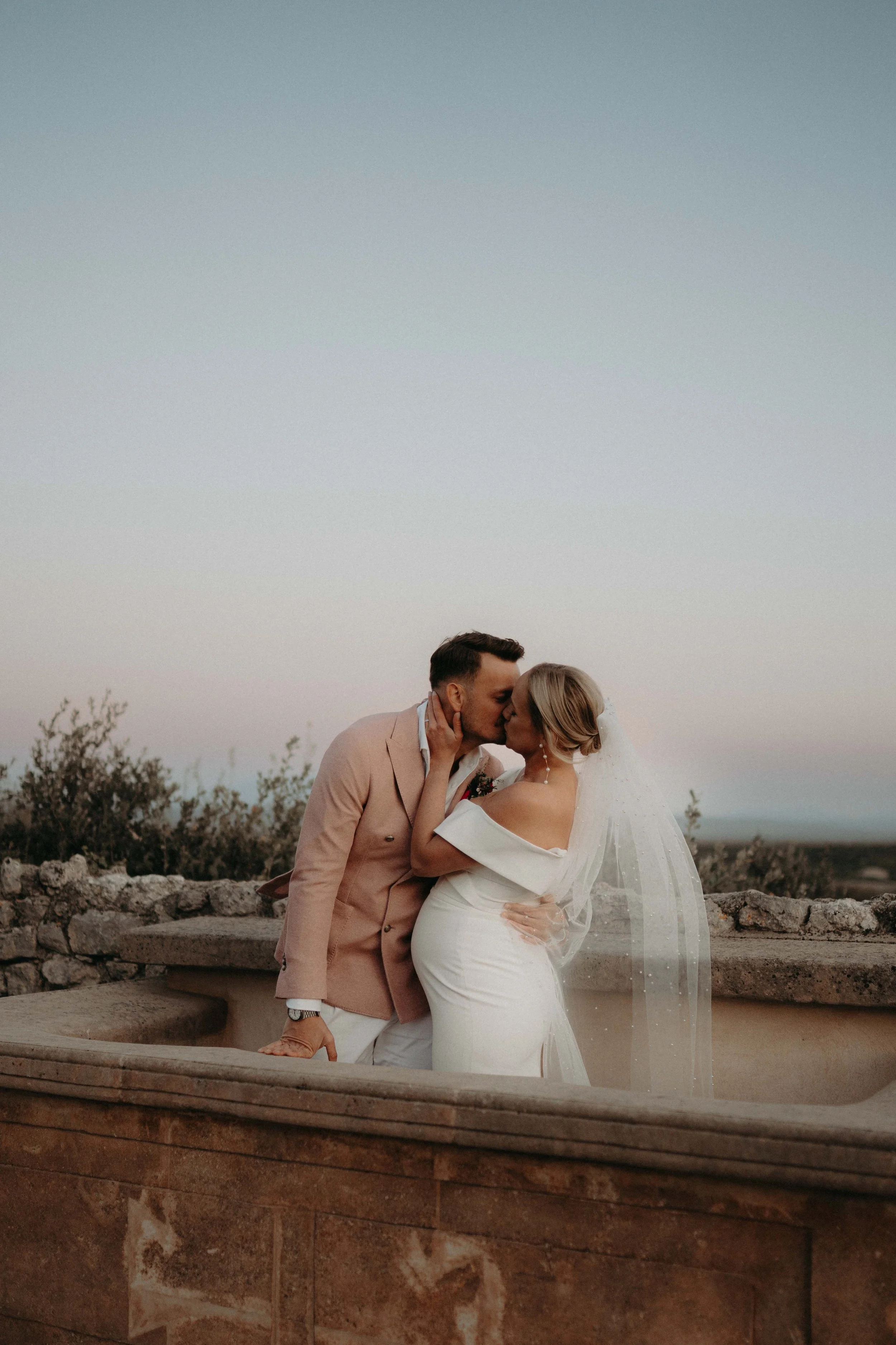 A couple dressed in wedding attire sharing a kiss outdoors during sunset, with a stone wall and sparse shrubbery in the background.