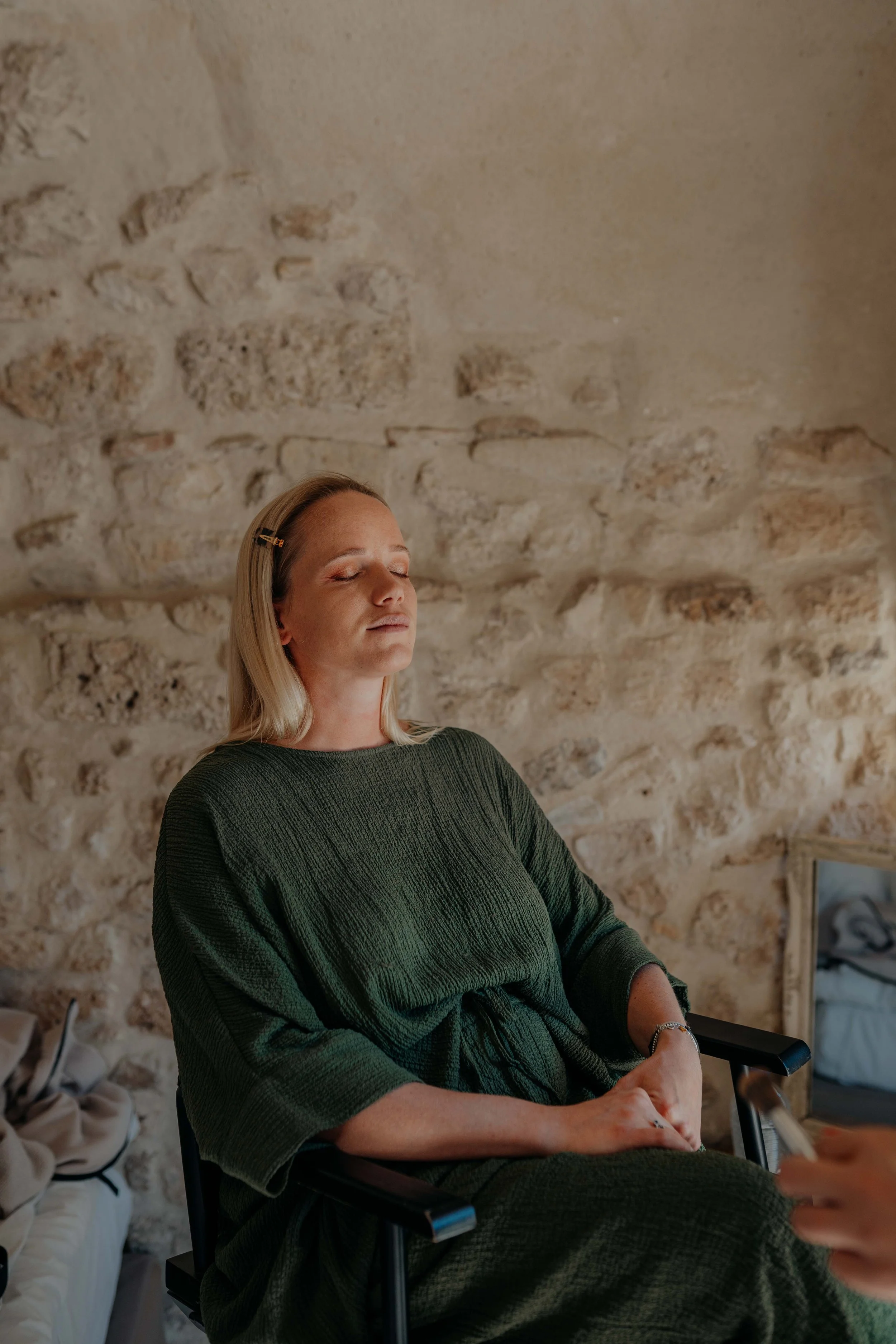 A woman with blonde hair, wearing a green textured dress, is sitting in a black chair with her eyes closed. She has a hair clip on one side of her hair and appears to be relaxed or meditating. The background features a rustic stone wall.