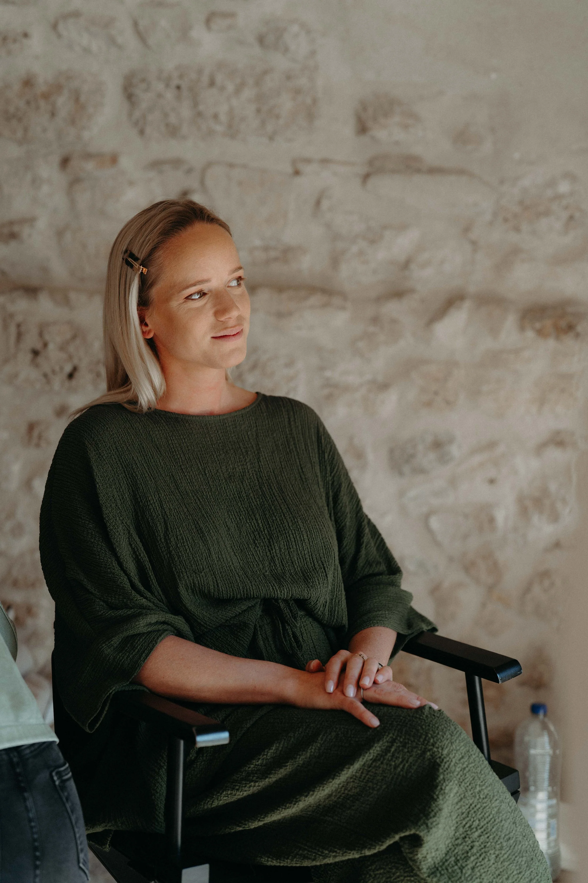 A woman with blonde hair, wearing a dark green textured dress, sitting on a black chair with her hands clasped, against a light stone wall background.