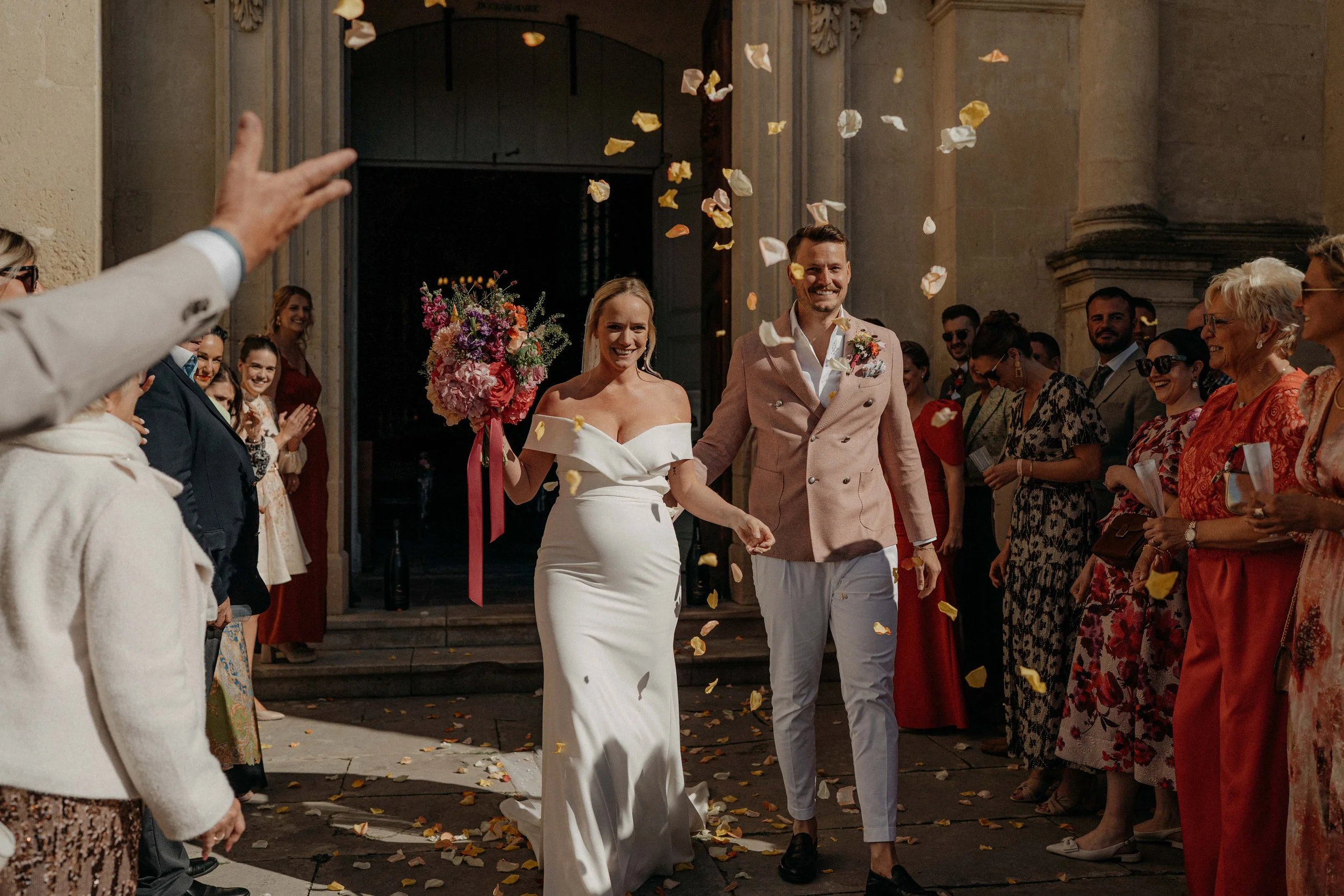 A newly married couple walks out of a church, smiling, as wedding guests throw flower petals around them. The bride is holding a bouquet of flowers and wearing a white dress, while the groom is dressed in a light pink blazer and white pants. Guests a