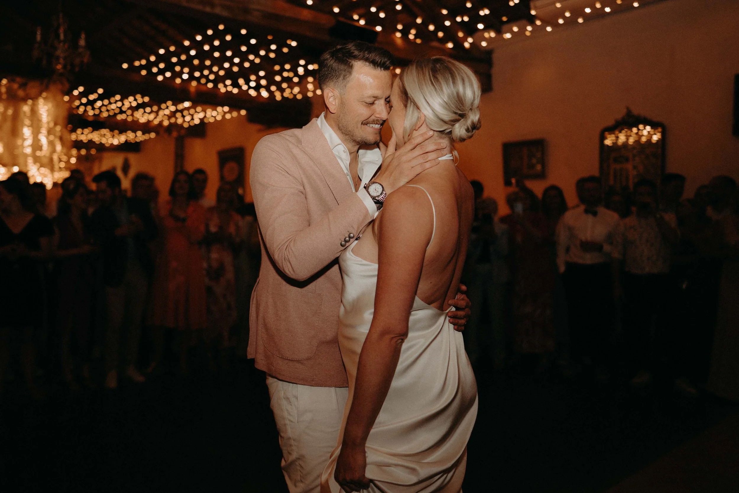 A couple sharing a dance at a wedding reception, with twinkling string lights overhead and other guests in the background.