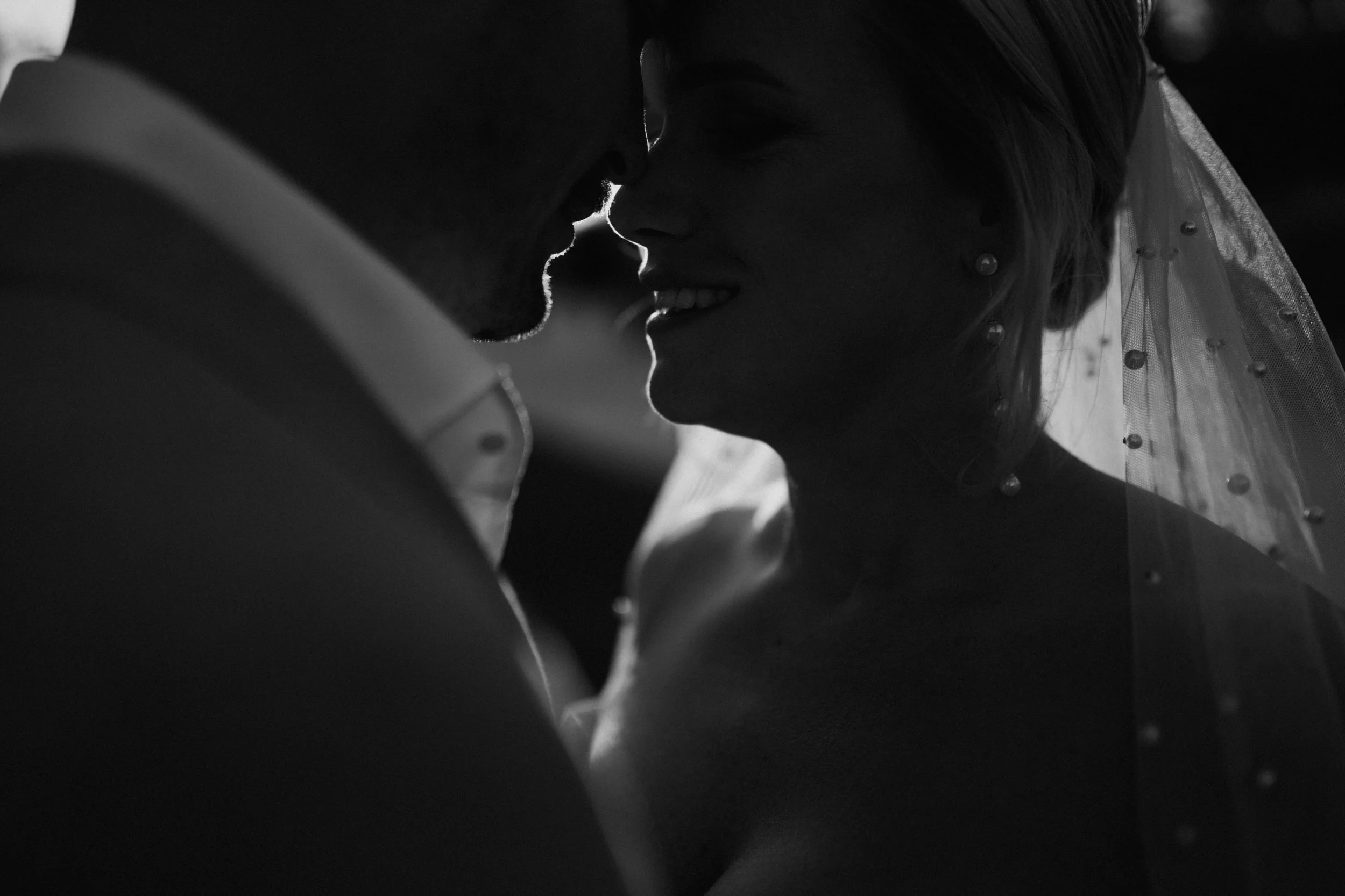 Black and white photo of a couple with their noses close together, sharing an intimate moment, possibly about to kiss. The woman is wearing a veil with small beads and earrings.
