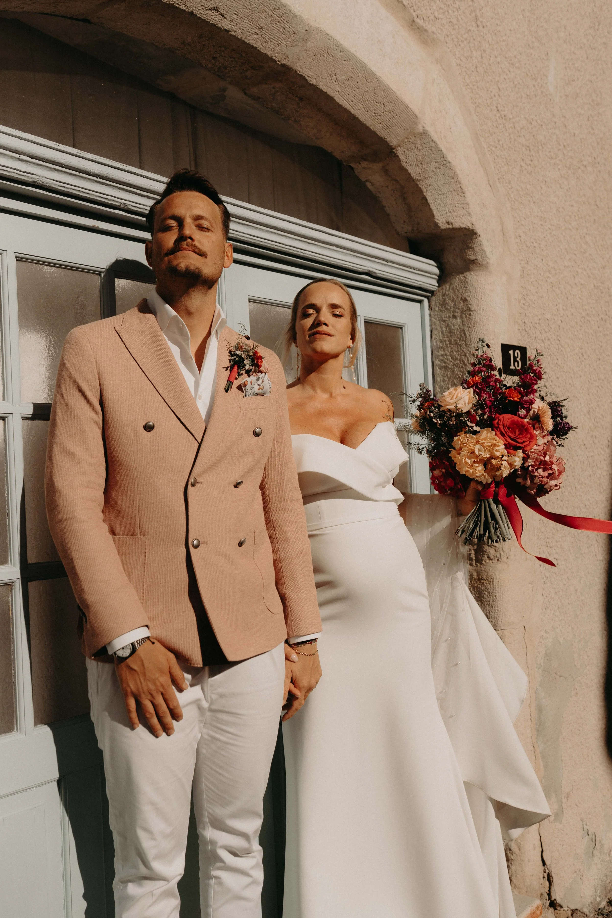 A man and a woman dressed in formal wedding attire, standing outdoors near a building with beige stone walls and a window, with a bouquet of flowers resting on his right arm.