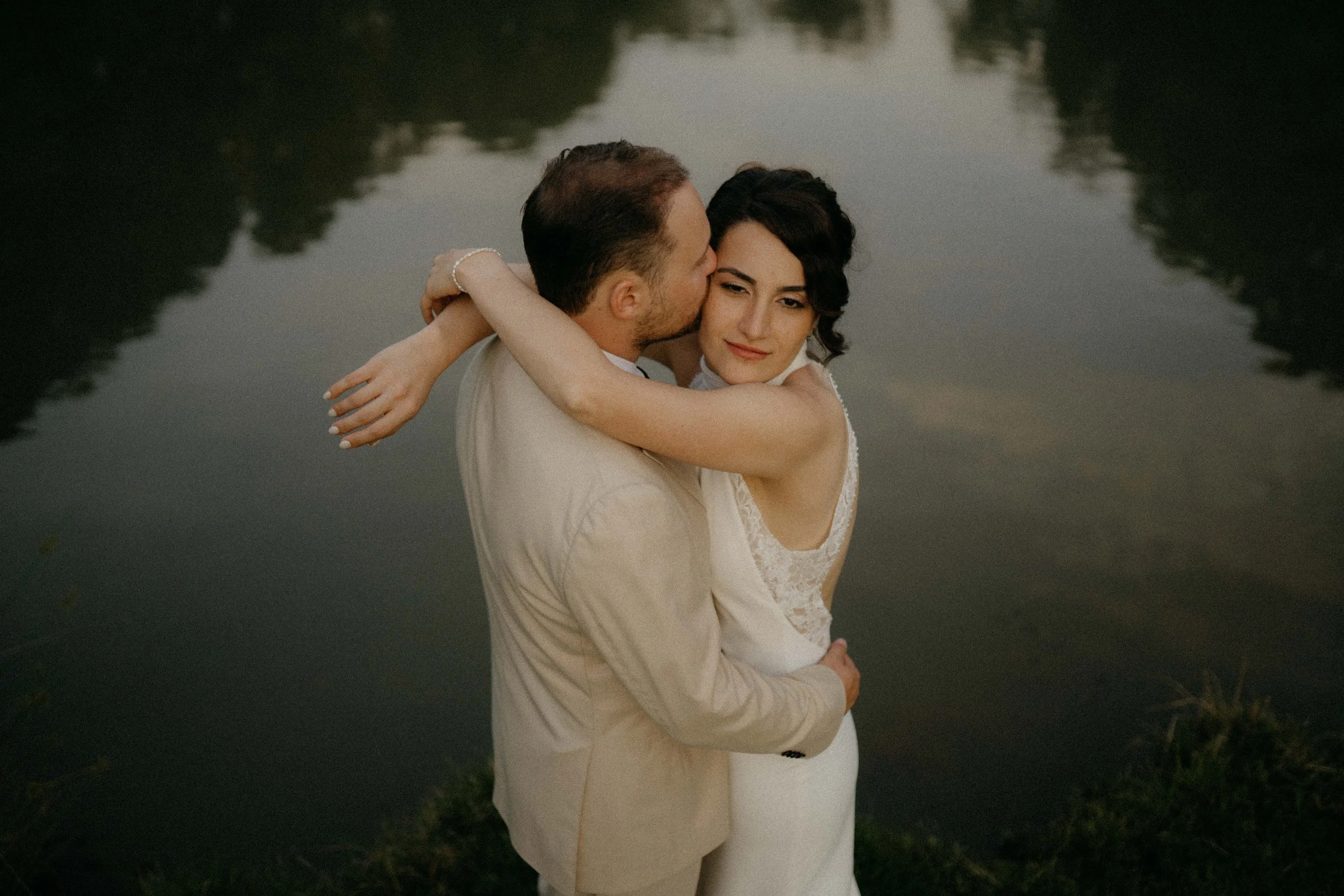 A couple embraces near a river at sunset, with both dressed in white; the man is kissing the woman's cheek while she looks at the camera.