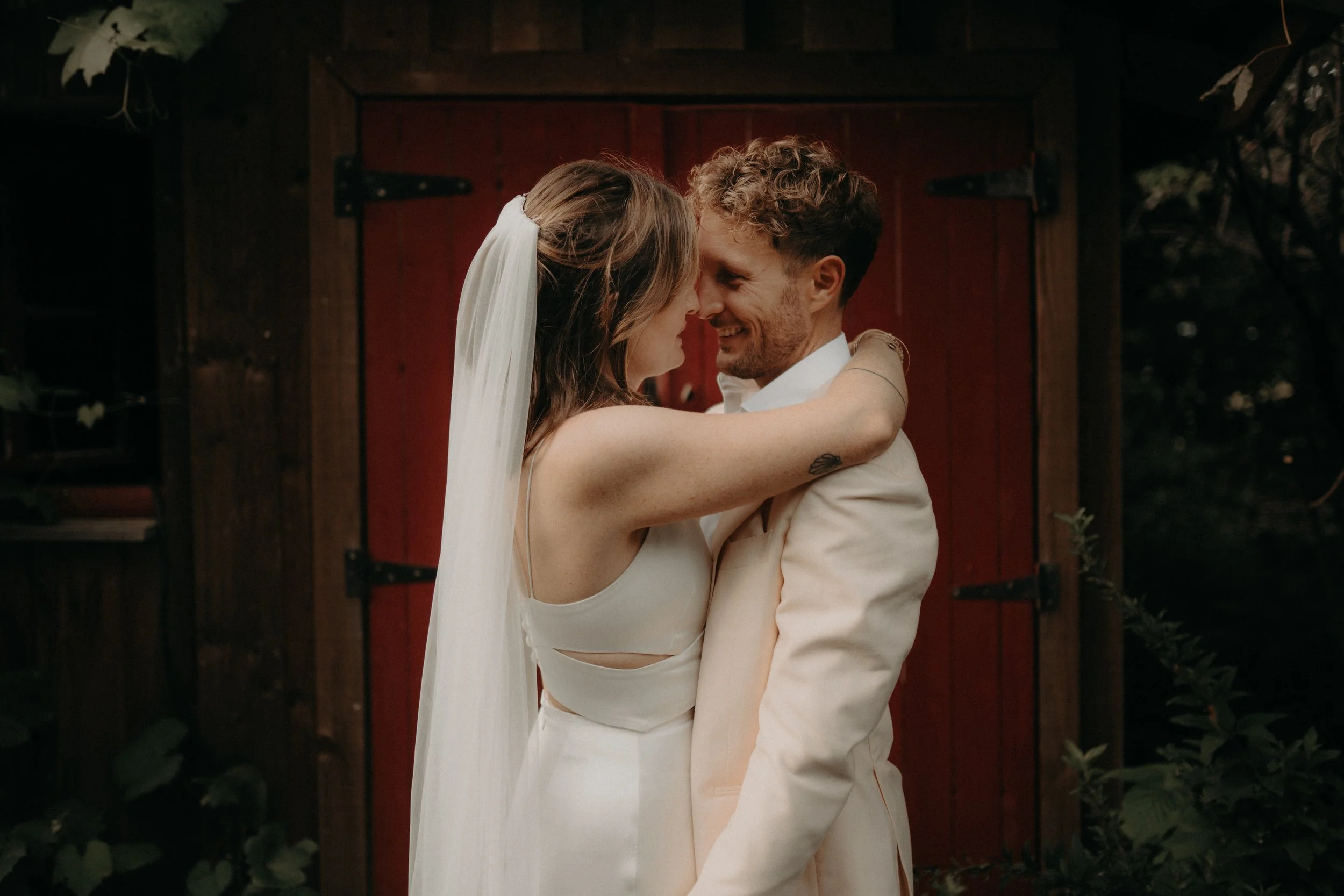 A couple in wedding attire embracing and touching foreheads in front of a red barn door, surrounded by greenery.