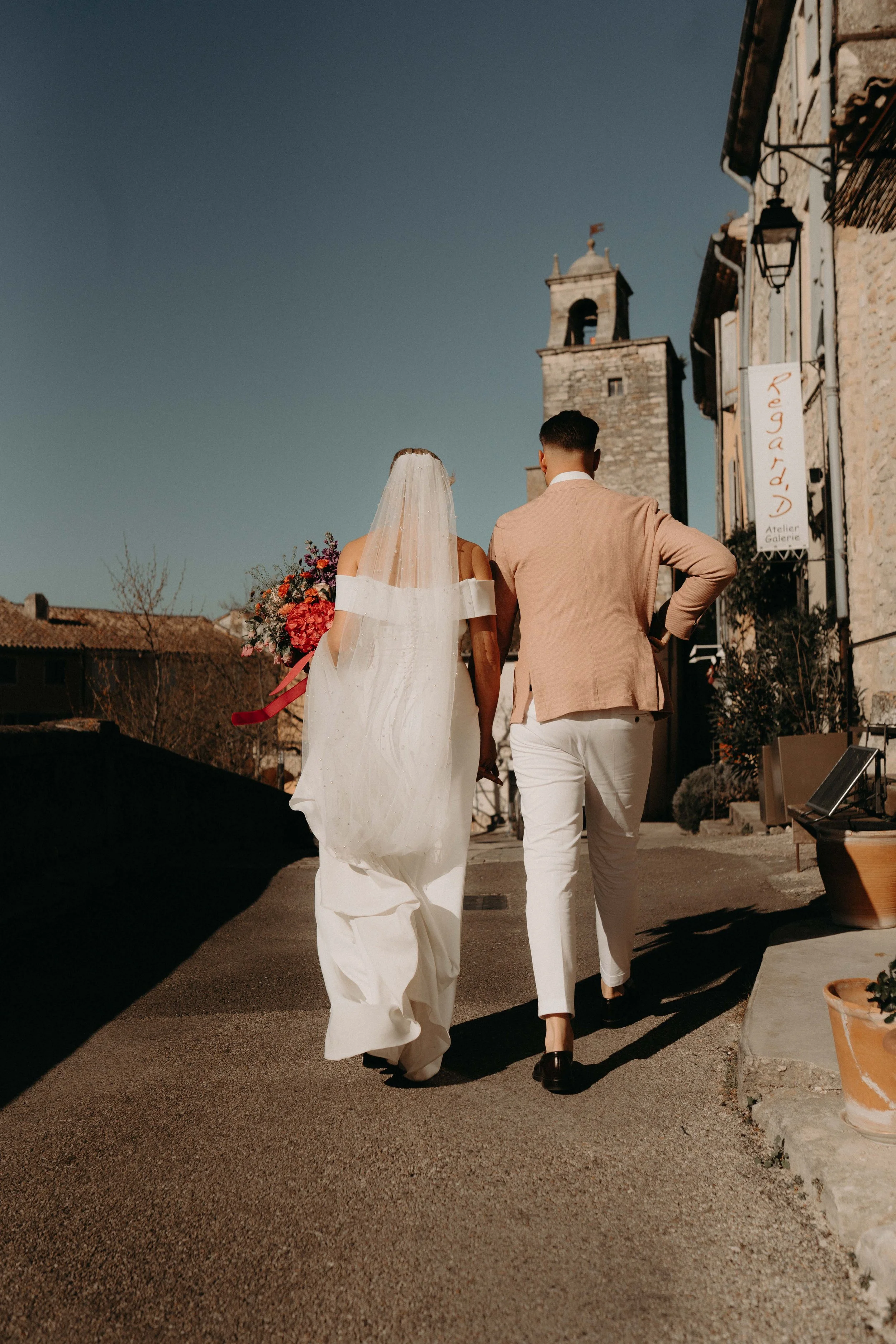 A bride in a white wedding gown and veil holding a bouquet walking with a groom in a beige suit along a street in a historic town with stone buildings and a tower in the background.