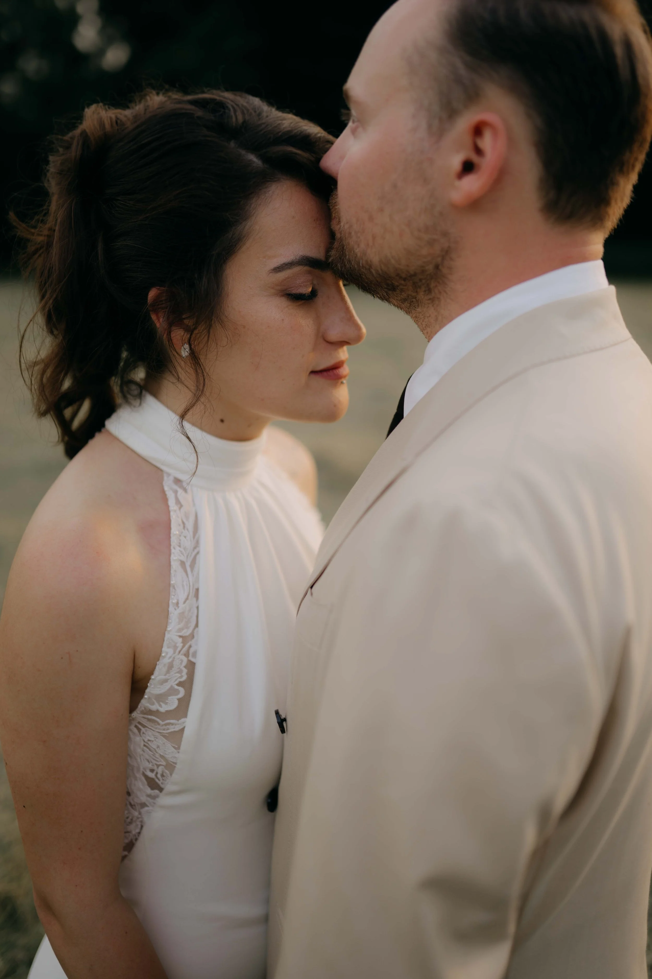 A close-up of a bride and groom close together with their foreheads touching during a wedding ceremony outdoors at sunset.
