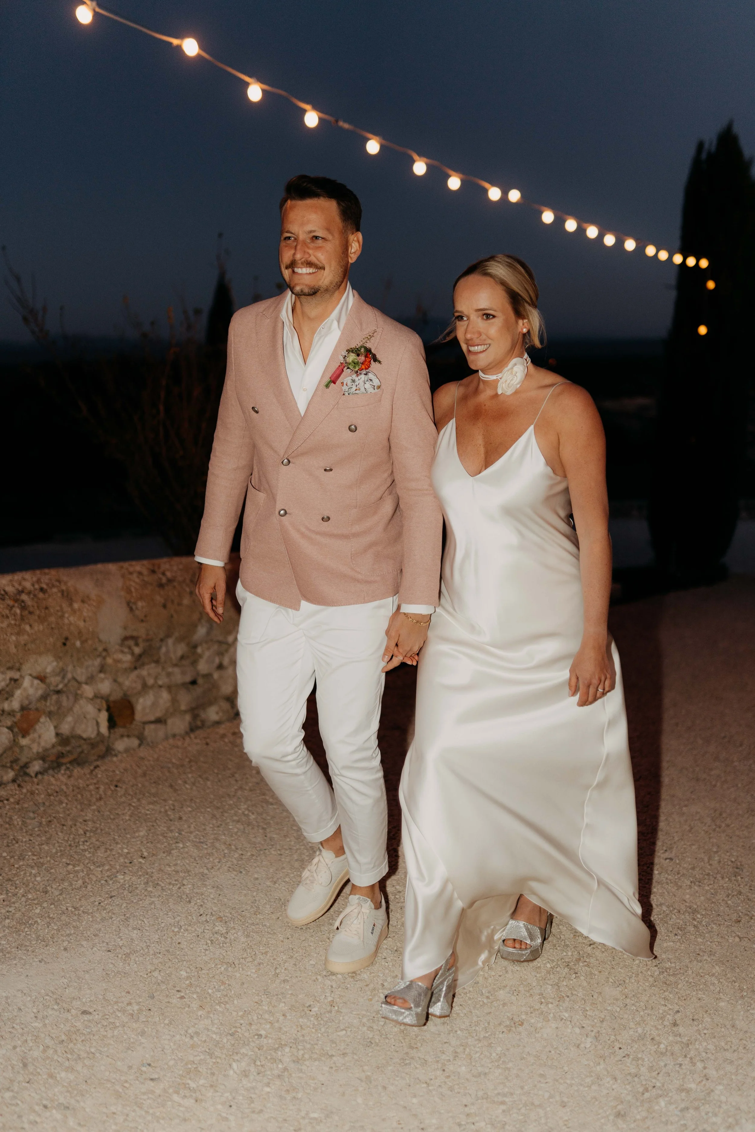 A couple walking hand in hand at night outdoors, with string lights overhead, the man wearing a pink blazer and white pants, and the woman in a white satin dress with silver high heels.