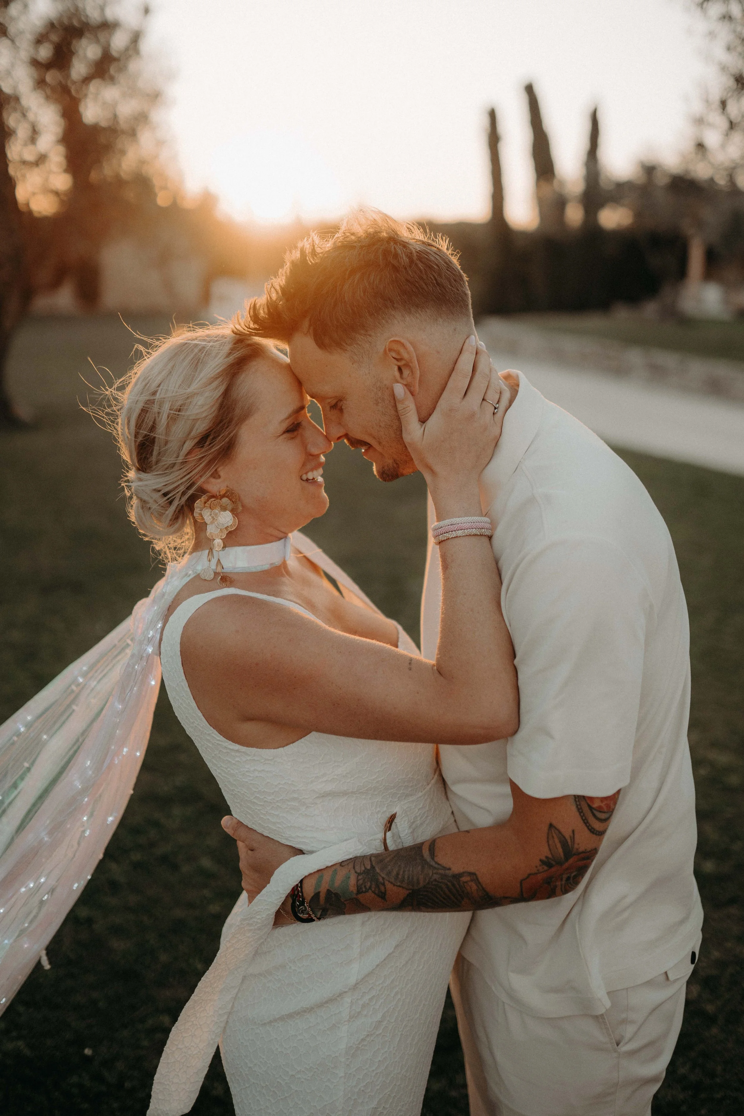 A couple during sunset, holding each other closely and touching foreheads, smiling, in an outdoor park setting.