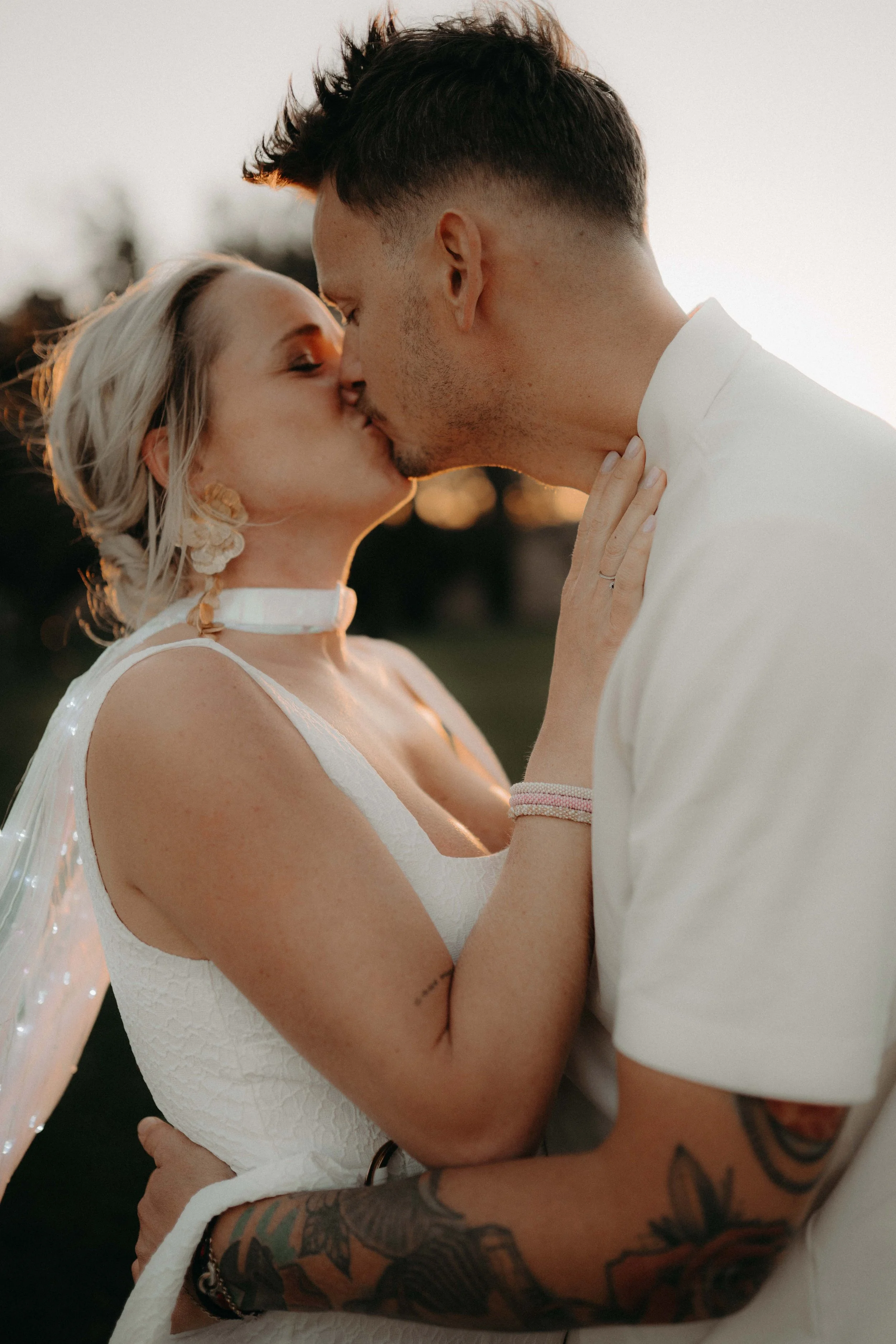 A close-up of a couple sharing a kiss, with the woman wearing a white dress and necklace, and the man wearing a white shirt, with tattooed arms visible. They are outdoors at sunset.