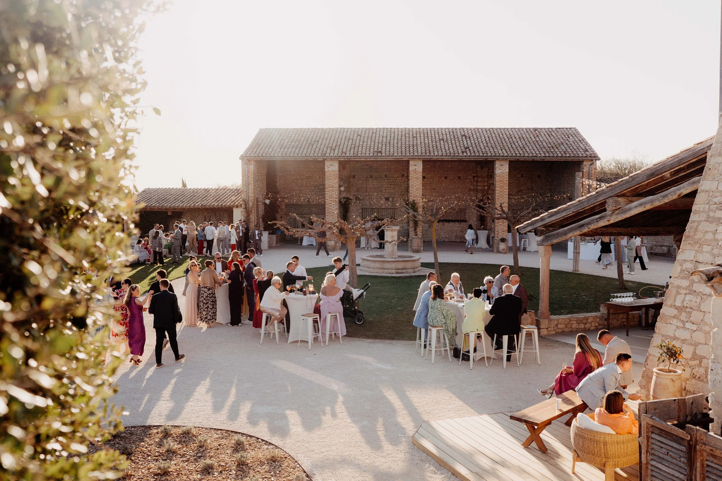 People gathered outdoors in a rustic courtyard during a social event or celebration, with tables, chairs, and a large brick building in the background.