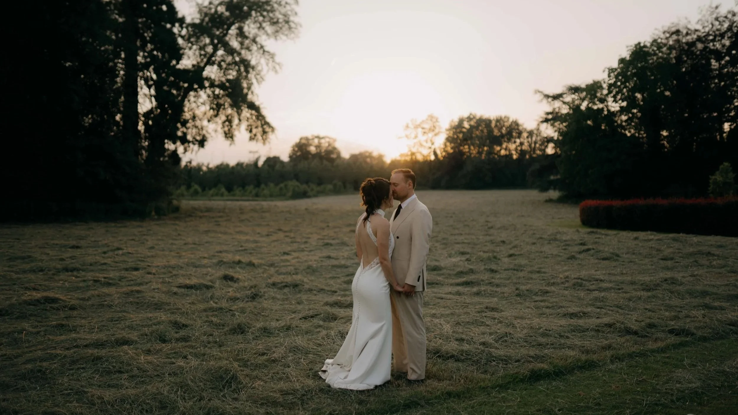 A bride and groom standing close together in a grassy field at sunset, holding hands and touching foreheads, with trees in the background.