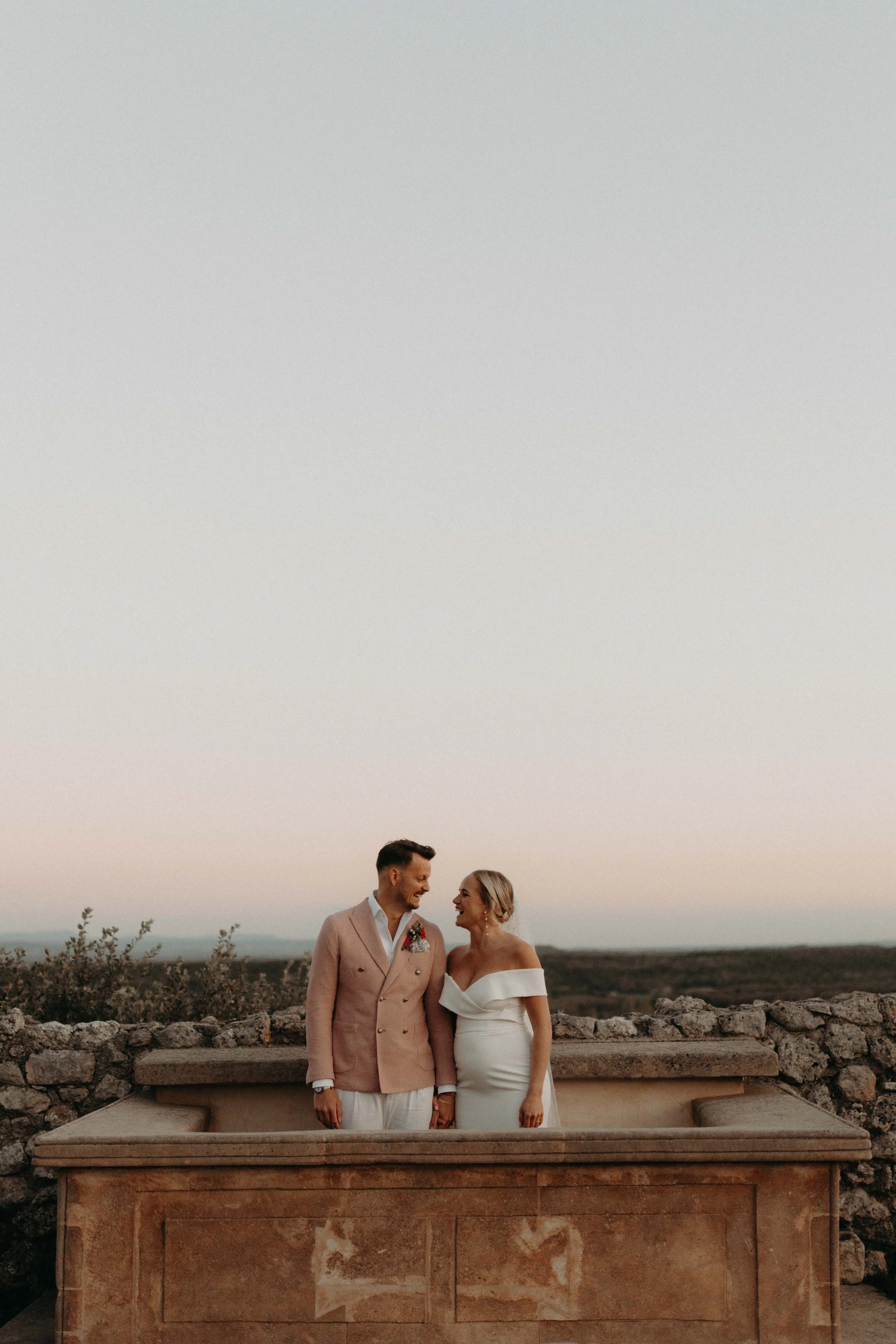 A newlywed couple standing outdoors on a stone platform, holding hands and smiling at each other, with a scenic landscape and a clear sky at sunset.