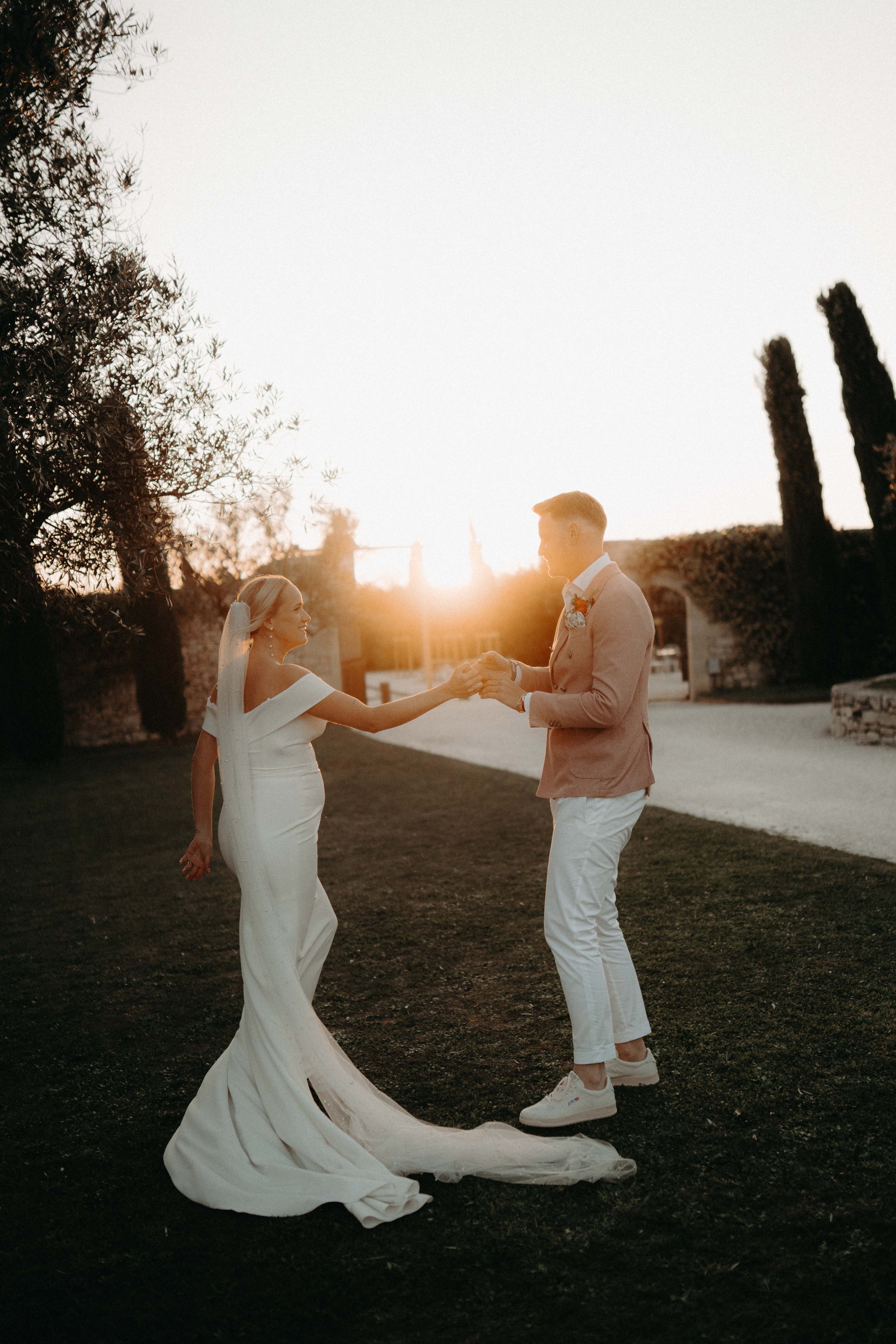 A bride and groom holding hands outdoors at sunset. The bride is wearing a white wedding dress and veil, and the groom is dressed in a light beige blazer, white pants, and sneakers. They are standing on grass, with trees and buildings in the backgrou