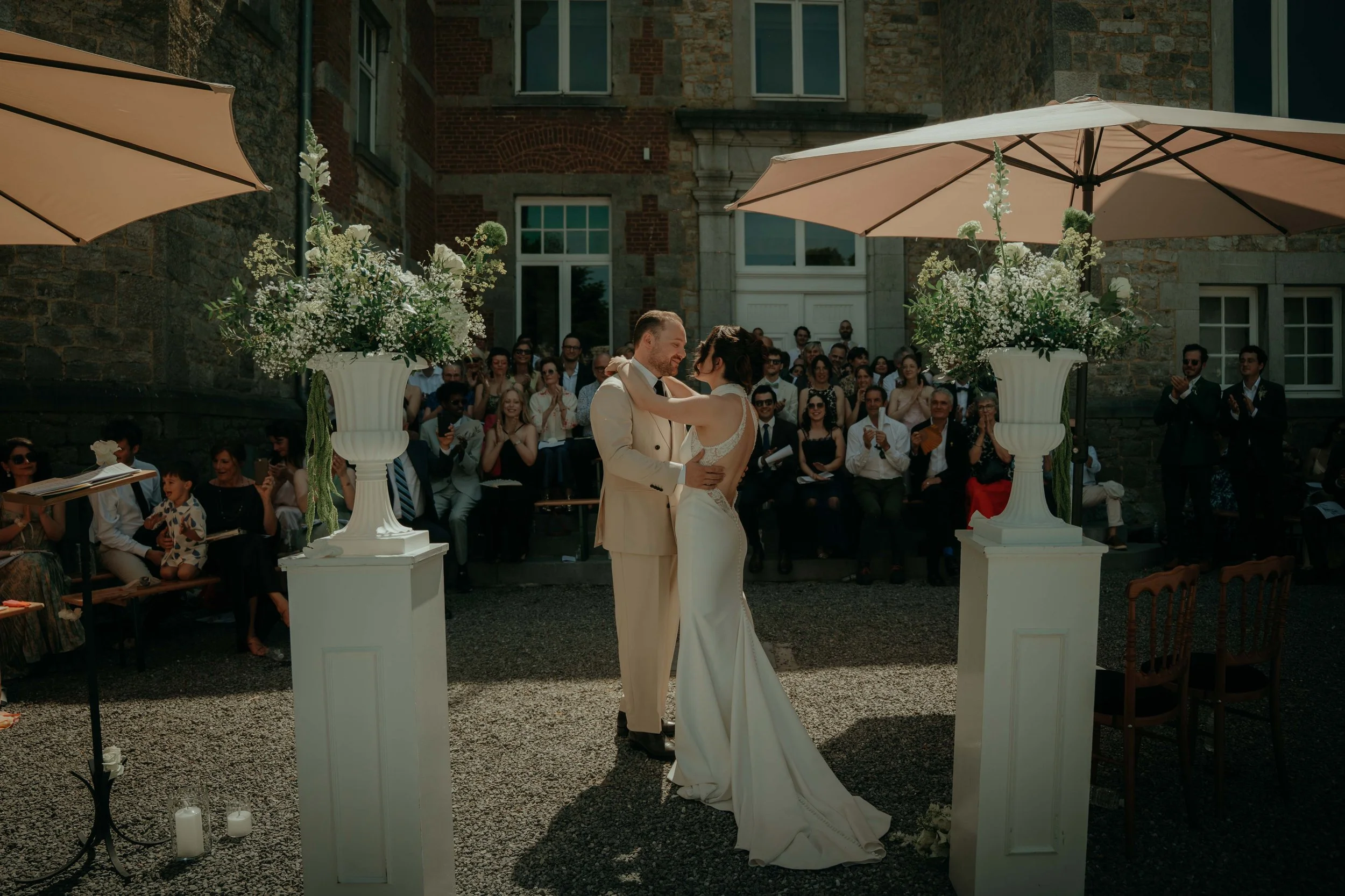 A couple exchanging wedding vows outdoors, with guests seated and standing behind them, during a wedding ceremony. The scene is set against a stone building with large windows and decorated with tall flower arrangements under umbrellas.