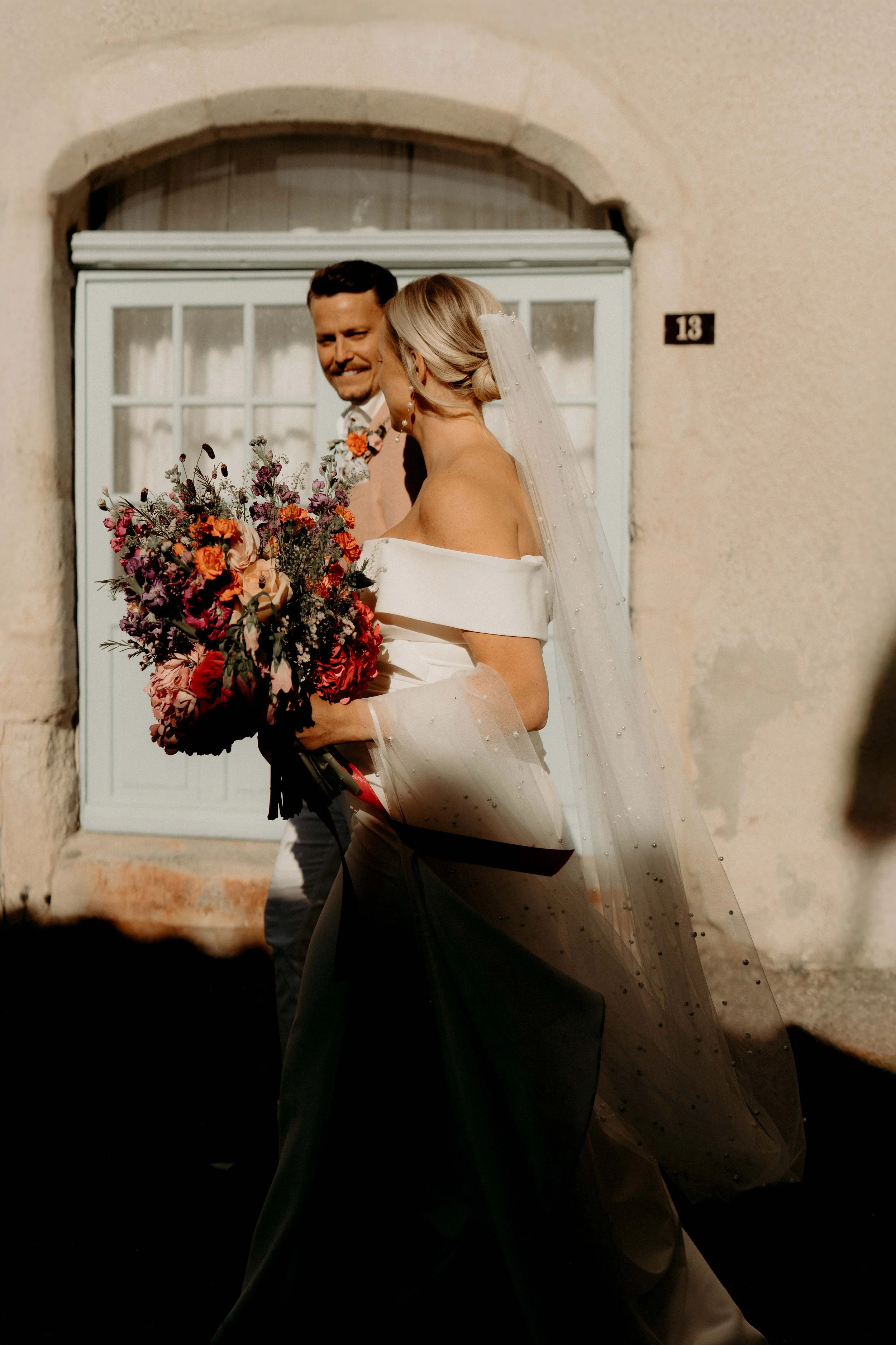A man and woman in wedding attire standing close together in front of a building with a stone arch and a window. The woman has a veil and is holding a large bouquet of colorful flowers. The man has short dark hair and a beard, smiling at the woman.