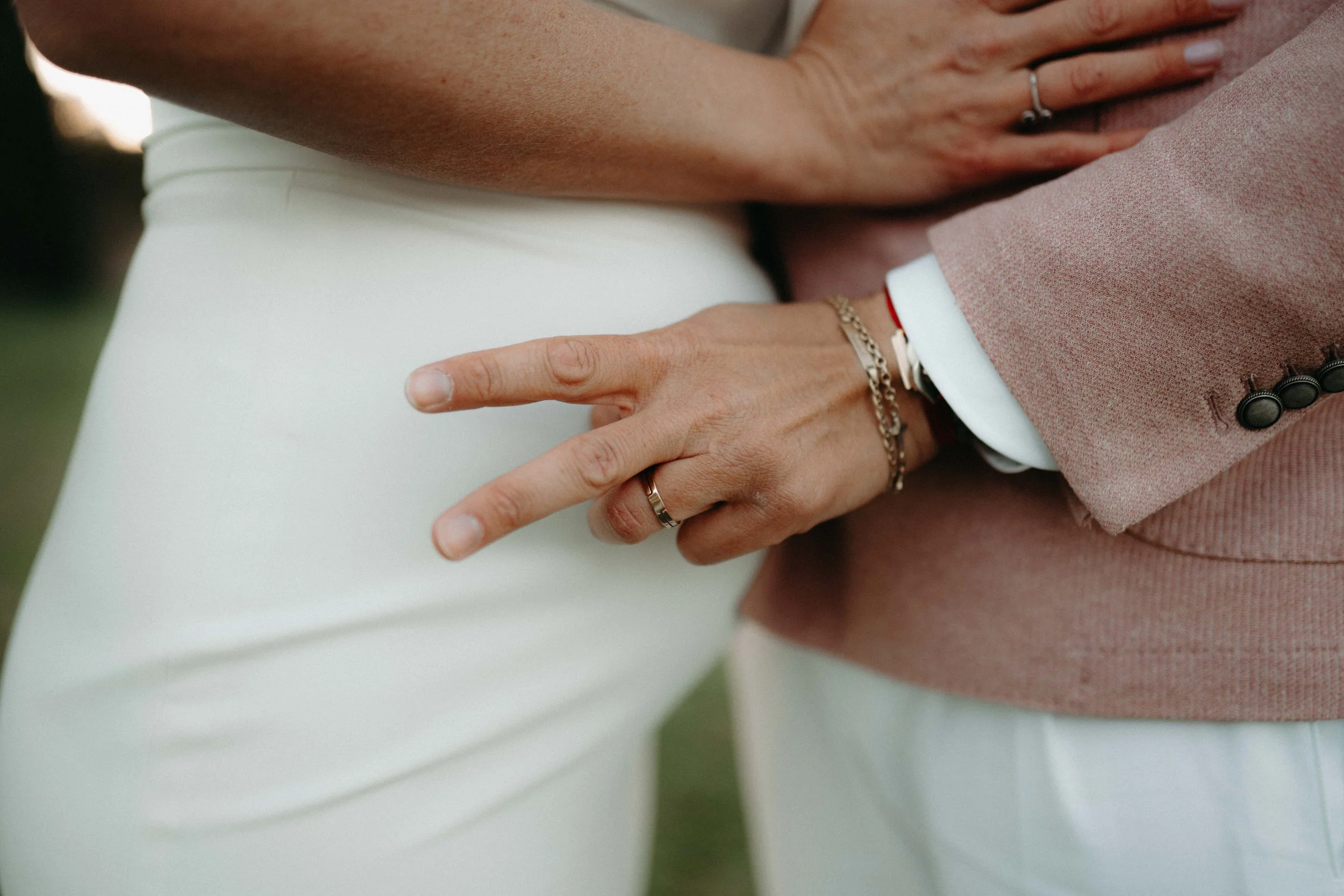 Close-up of a person in a pink suit and white shirt, resting their hand on a white surface. The person is wearing rings and a bracelet, and the hand is touching another person's hand.
