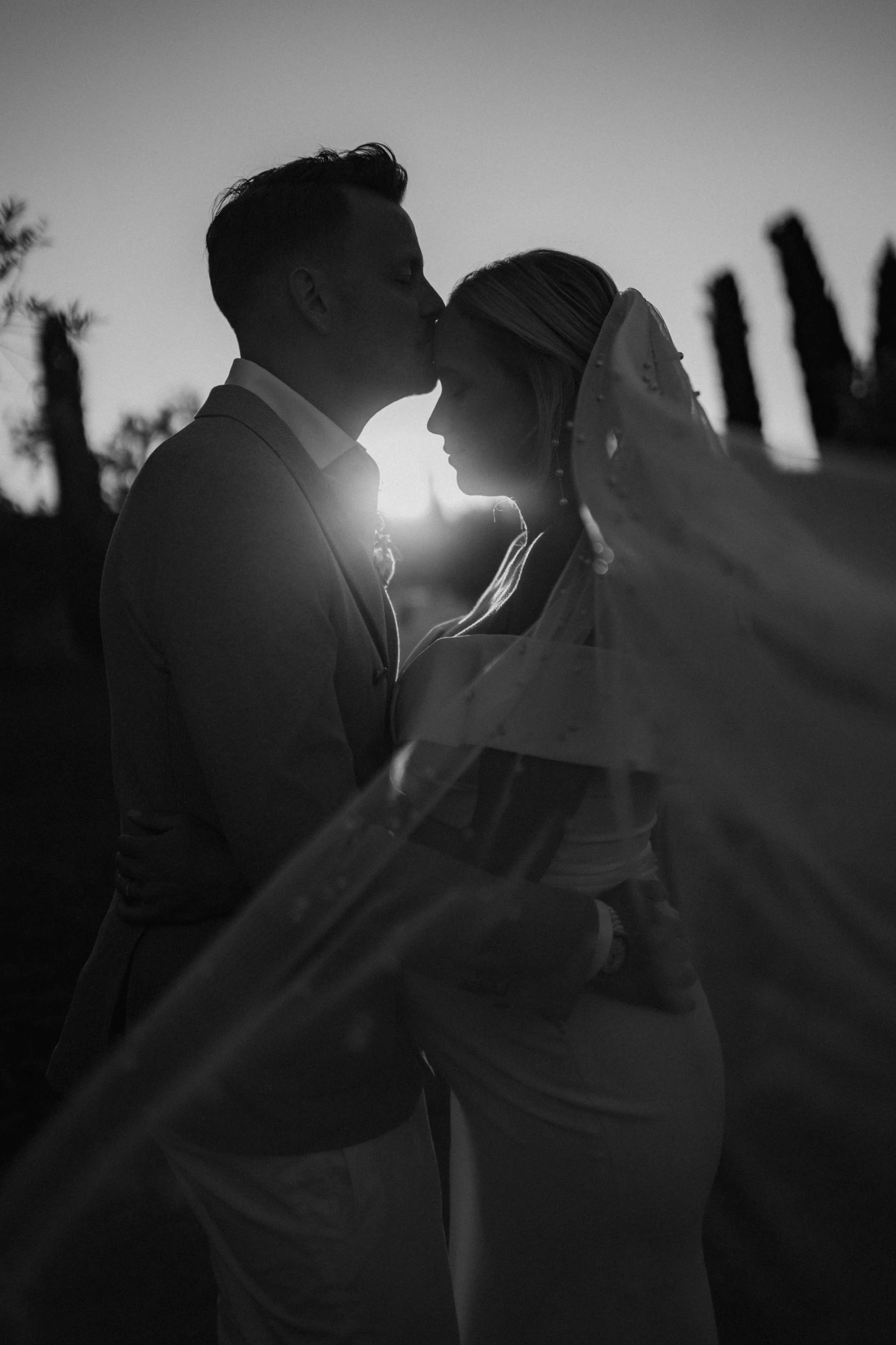 A black and white photo of a couple, with the man kissing the woman's forehead during sunset. The man is wearing a suit and the woman is in a wedding dress, with a veil. The background shows trees and a clear sky.