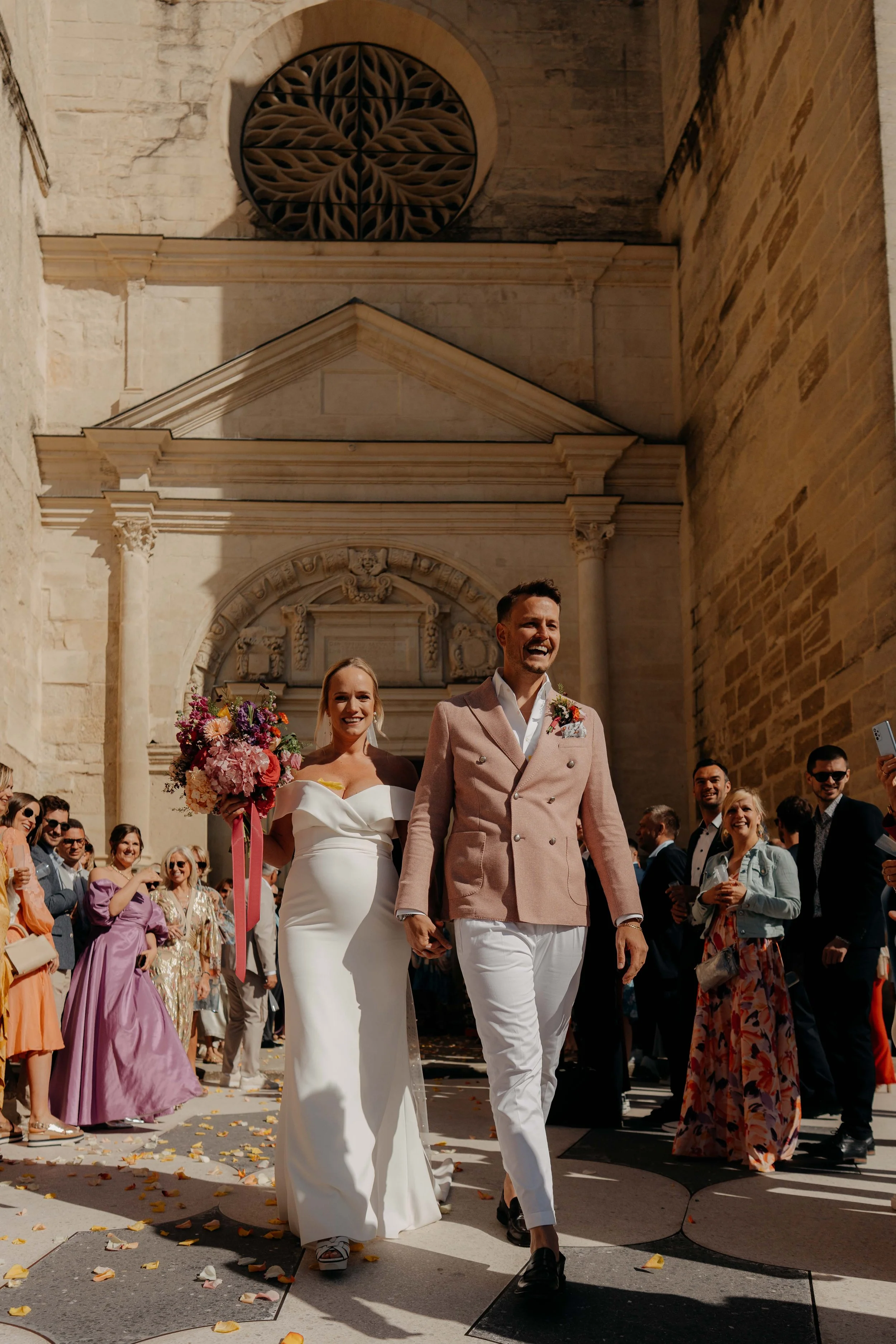A newlywed couple walking outdoors on a sunny day, surrounded by friends and family, in front of a historic building with stone architecture, celebration atmosphere, flower bouquets, and guests dressed in colorful formal attire.