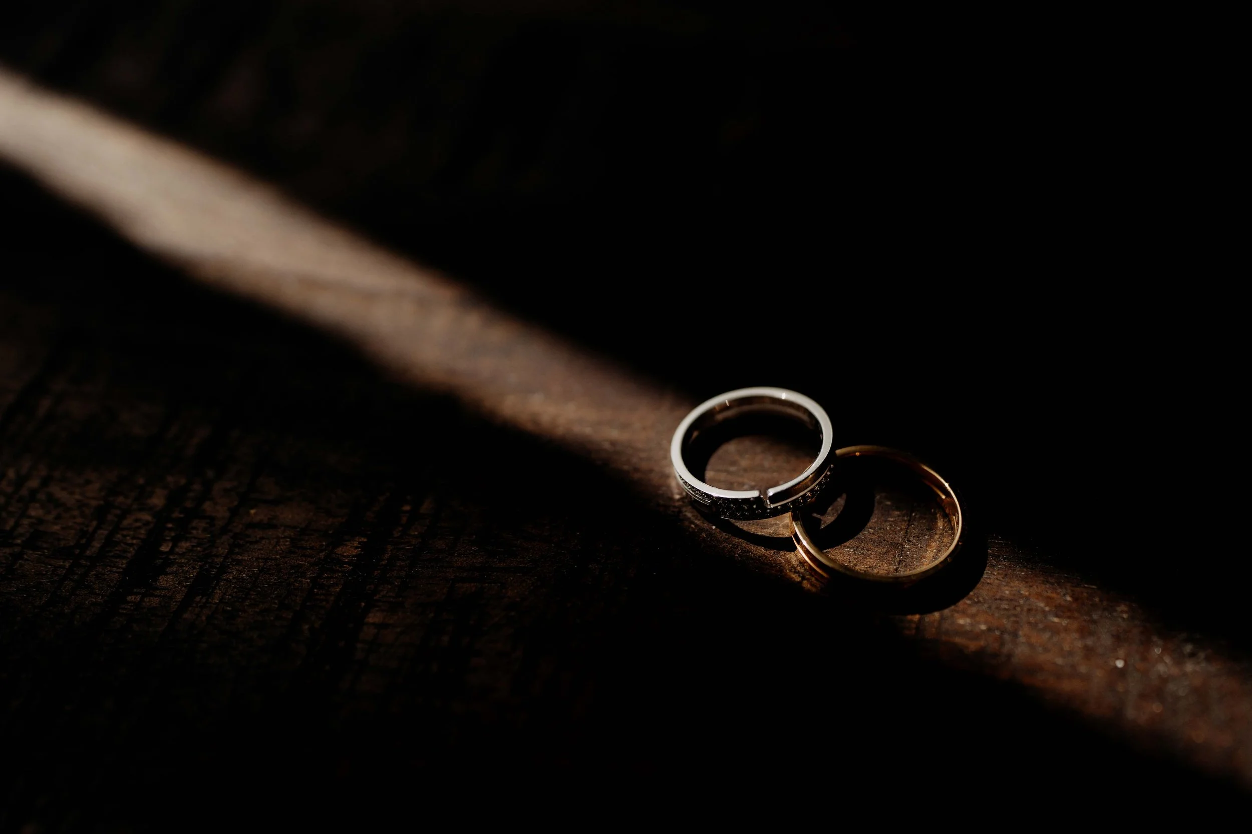 Two wedding rings, one silver and one gold, resting on a dark wooden surface with dramatic lighting.