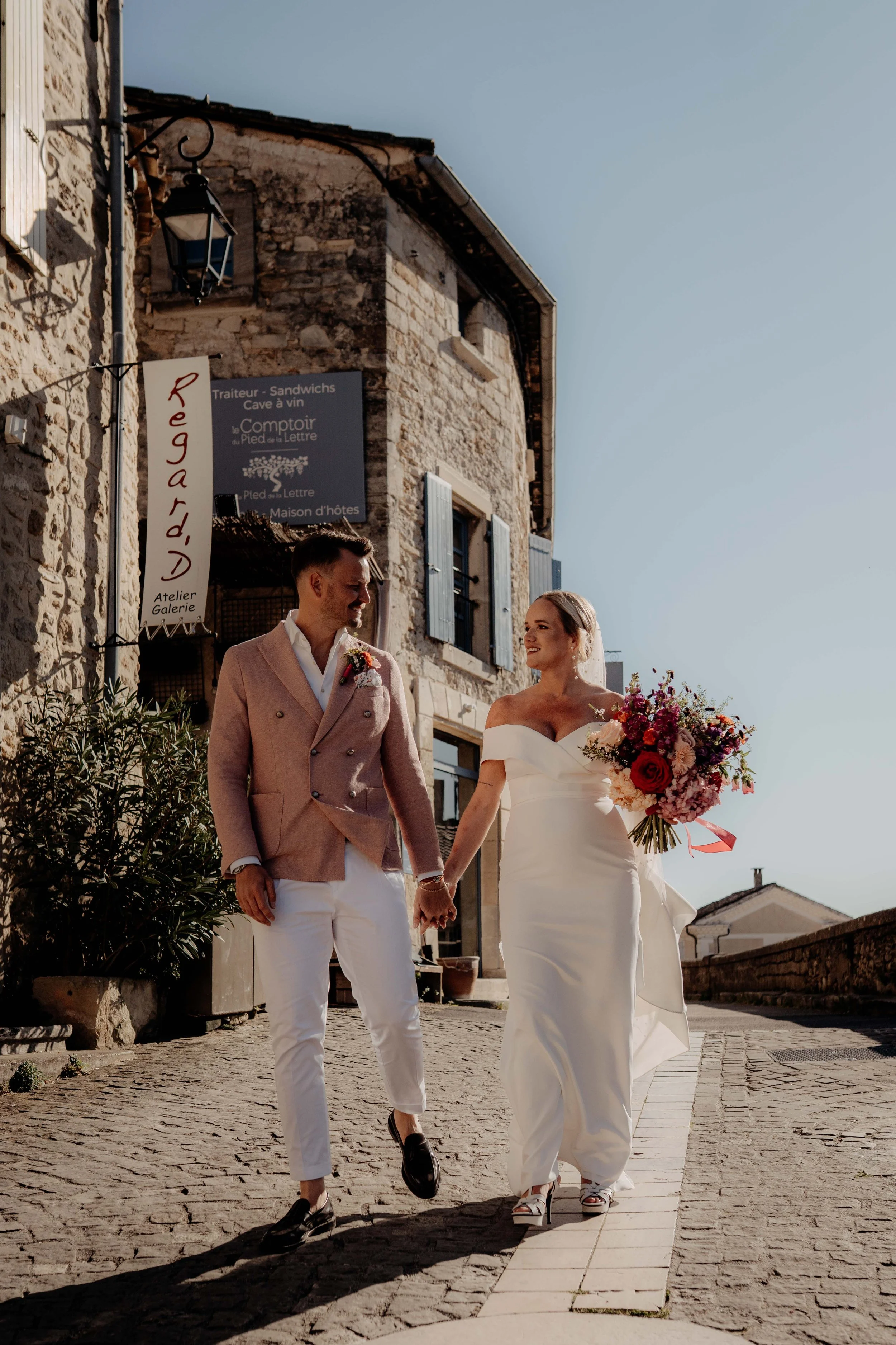 A bride and groom walking hand in hand down a cobblestone street, holding hands. The bride is wearing a white off-the-shoulder wedding gown and heels, holding a large colorful bouquet. The groom is dressed in a light pink blazer, white shirt, and whi