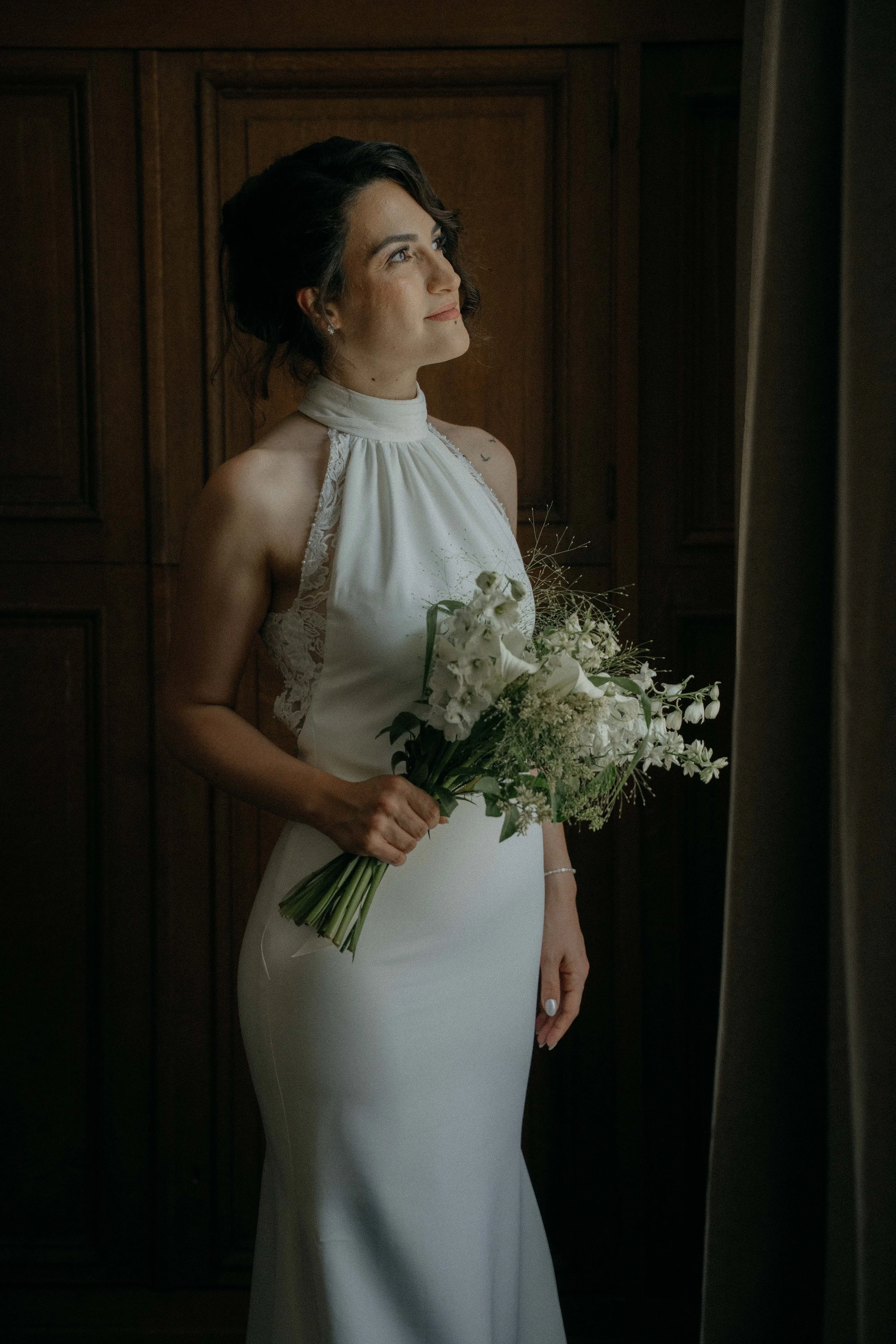 A woman dressed in a white gown holding a bouquet of white flowers, standing indoors against a wooden wall.