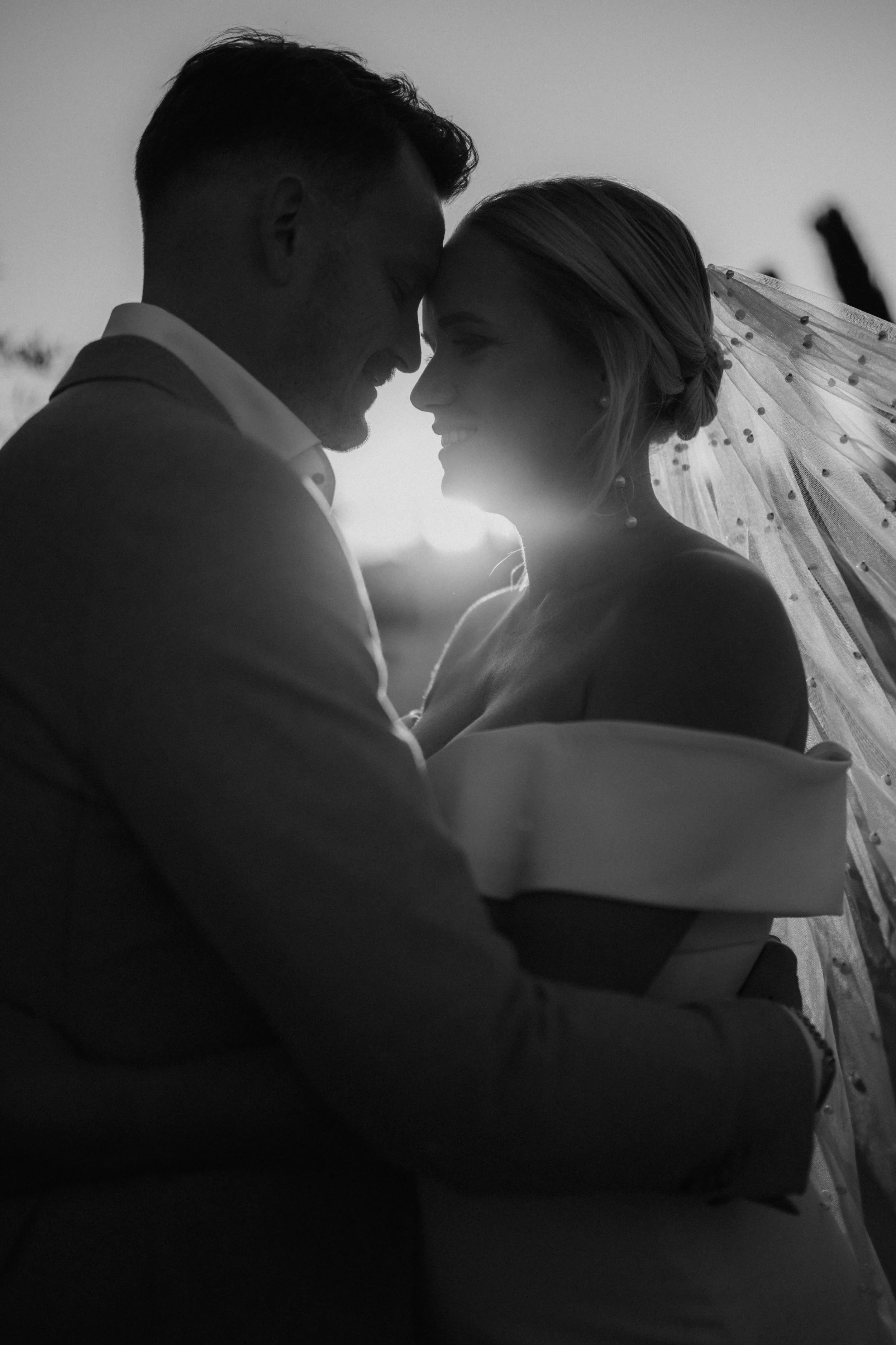 A black and white photo of a couple close together, smiling, with their foreheads touching. The woman is wearing a veil with small beads, indicating a wedding.