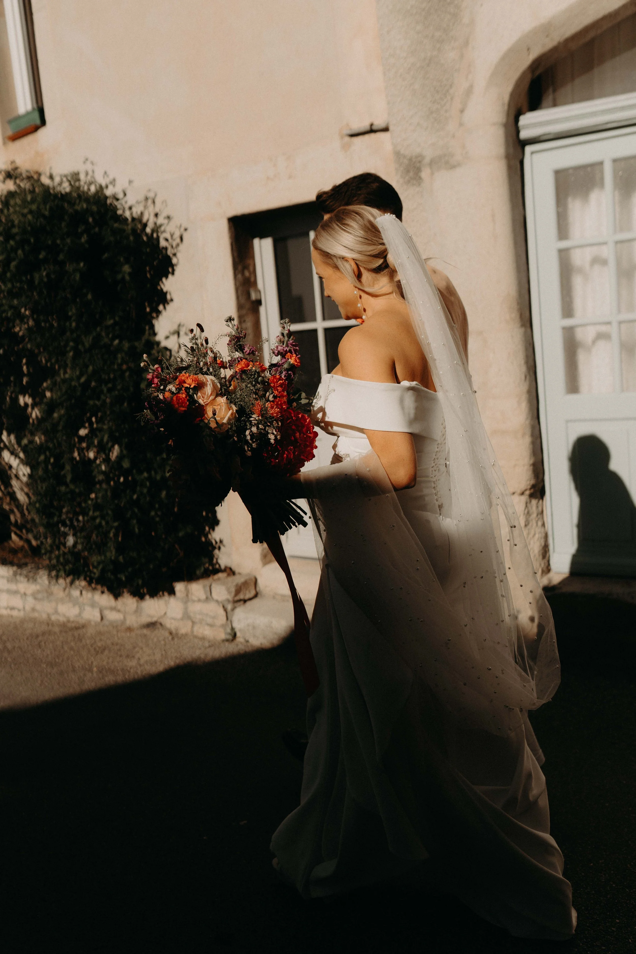 A bride with blonde hair in an updo, wearing a white wedding gown and veil, holding a large colorful bouquet, smiling and leaning toward a groom, outside a stone building with windows and a bush.