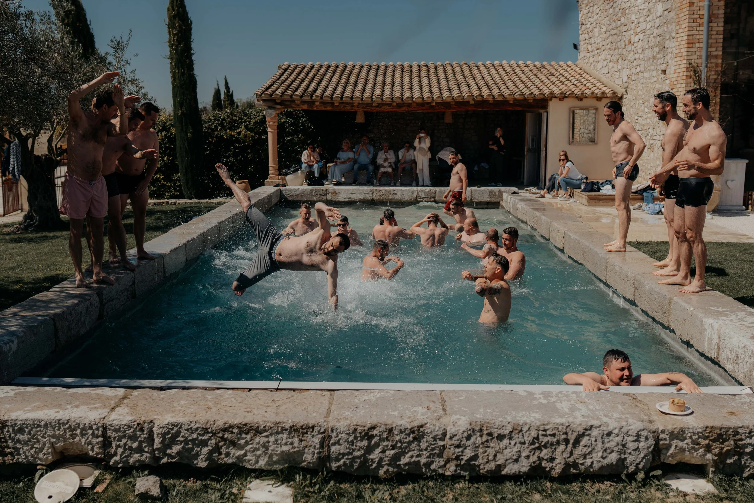People enjoying a pool party at a rustic outdoor setting with a stone building and a covered area, some standing on the pool's edge and others swimming in the water.