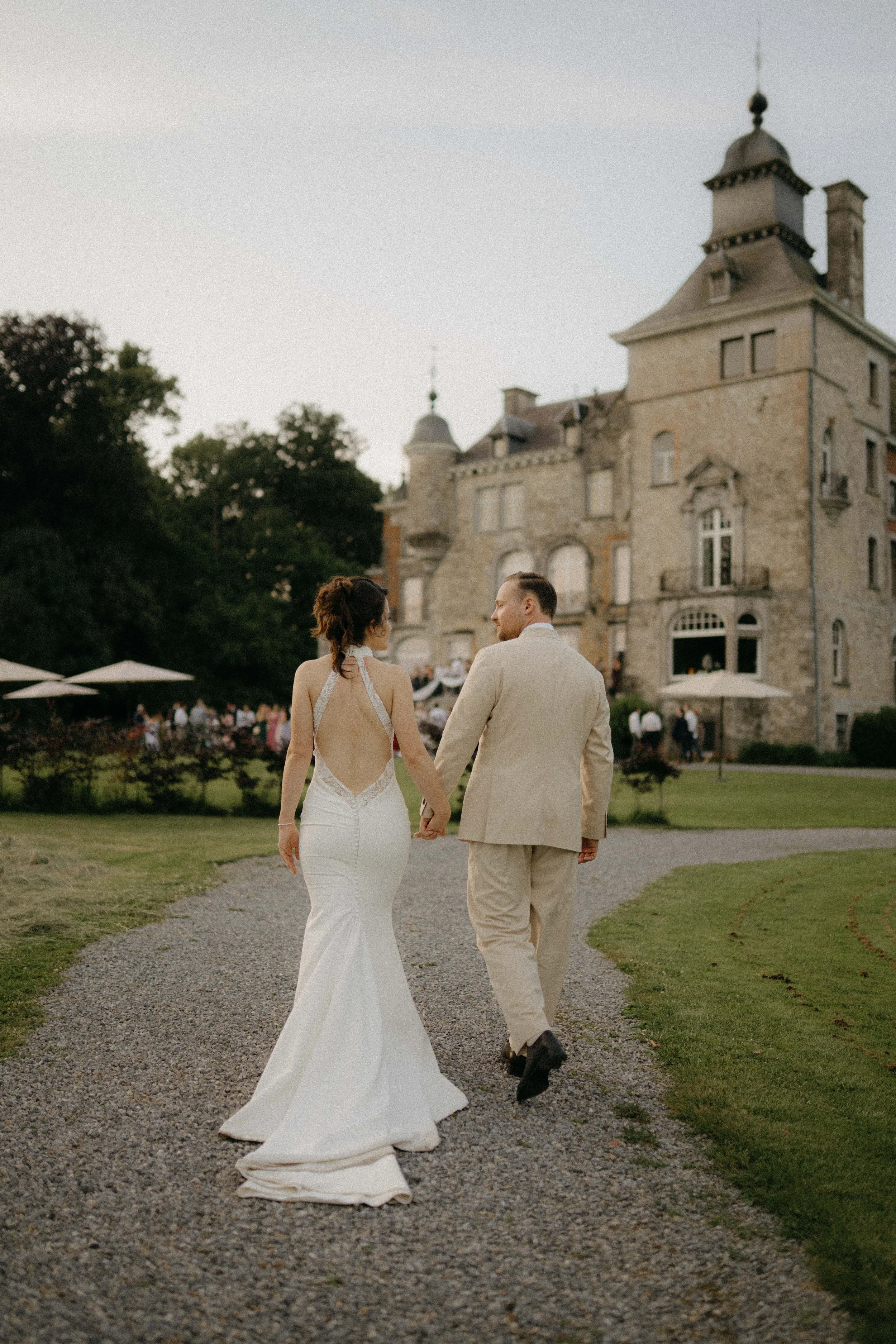 A bride and groom holding hands and walking away from the camera on a gravel pathway in front of a large historic stone mansion with a tower. The bride is wearing a white wedding dress with a low back and the groom is dressed in a light beige suit. T