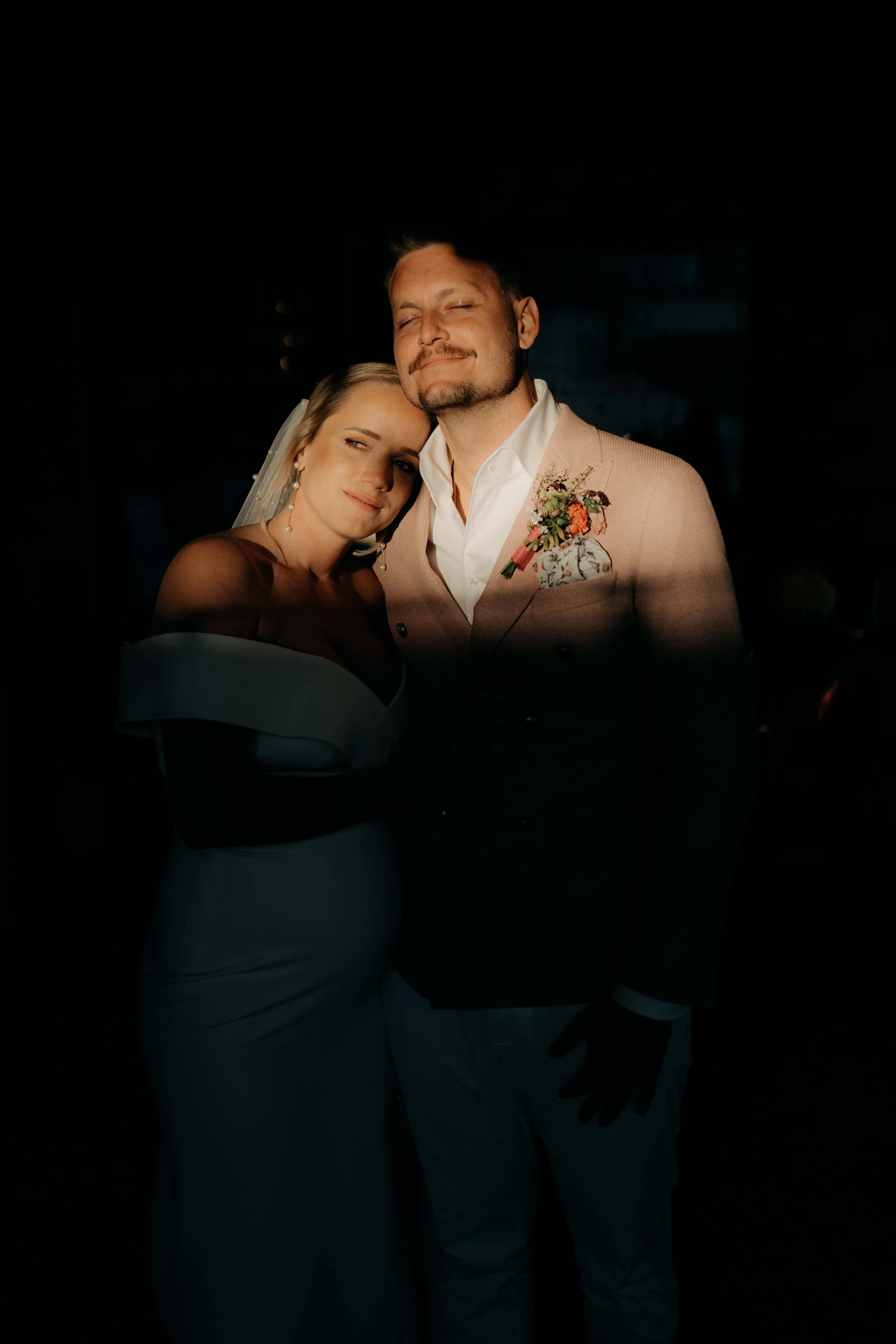 A bride and groom embracing at their wedding, smiling with eyes closed, in a dark background.