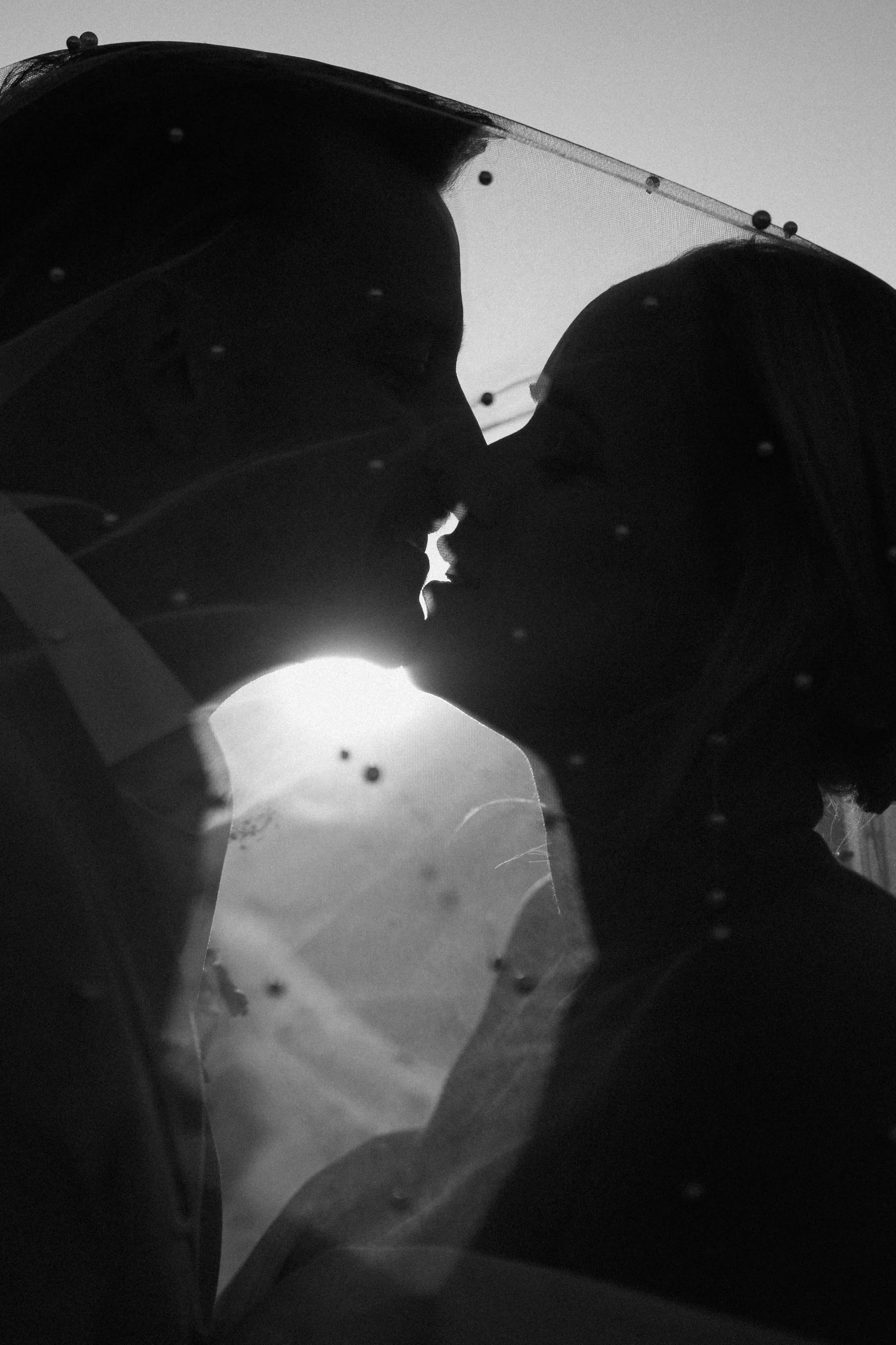 Black and white photo of two people in close proximity, with their faces nearly touching, framed by the silhouette of an umbrella with raindrops on it. The background is bright, possibly the sky, creating a contrast with the dark figures.