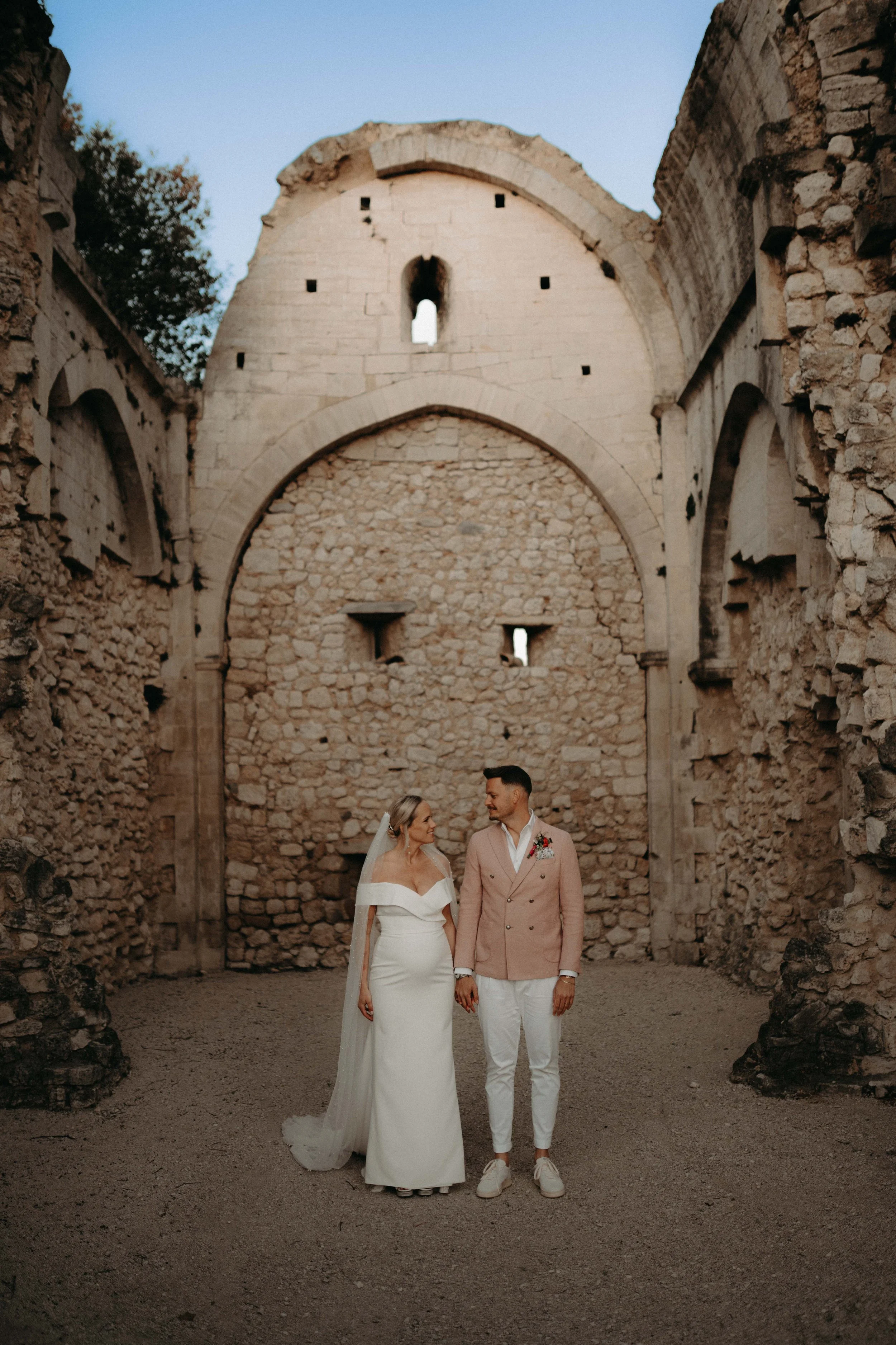 A bride and groom holding hands and walking through ancient stone ruins, with arches and windows in the background, at sunset.