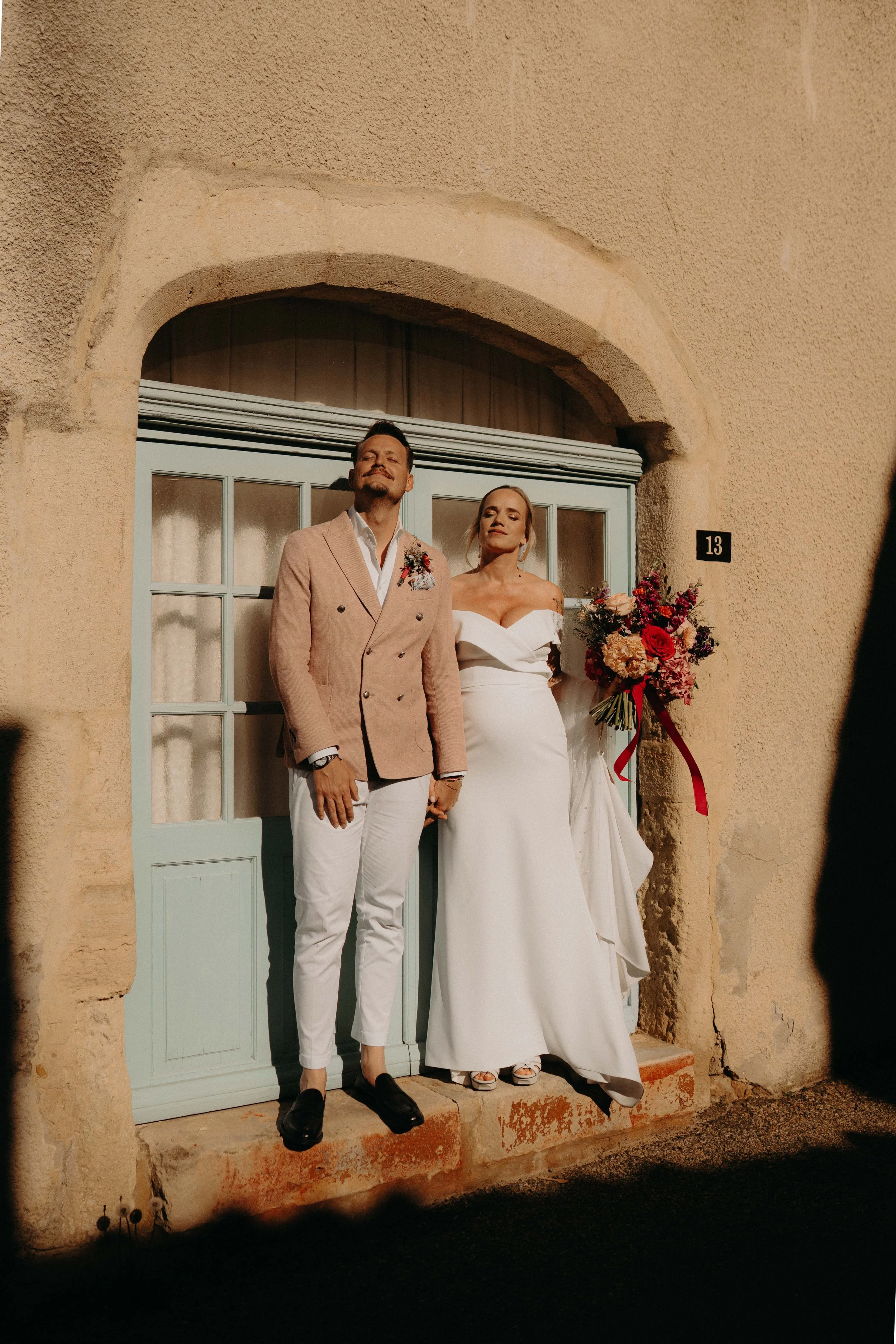 A man and woman standing outside a building, dressed in wedding attire, with the man in a light-colored blazer and the woman in a white wedding gown holding a bouquet of flowers.