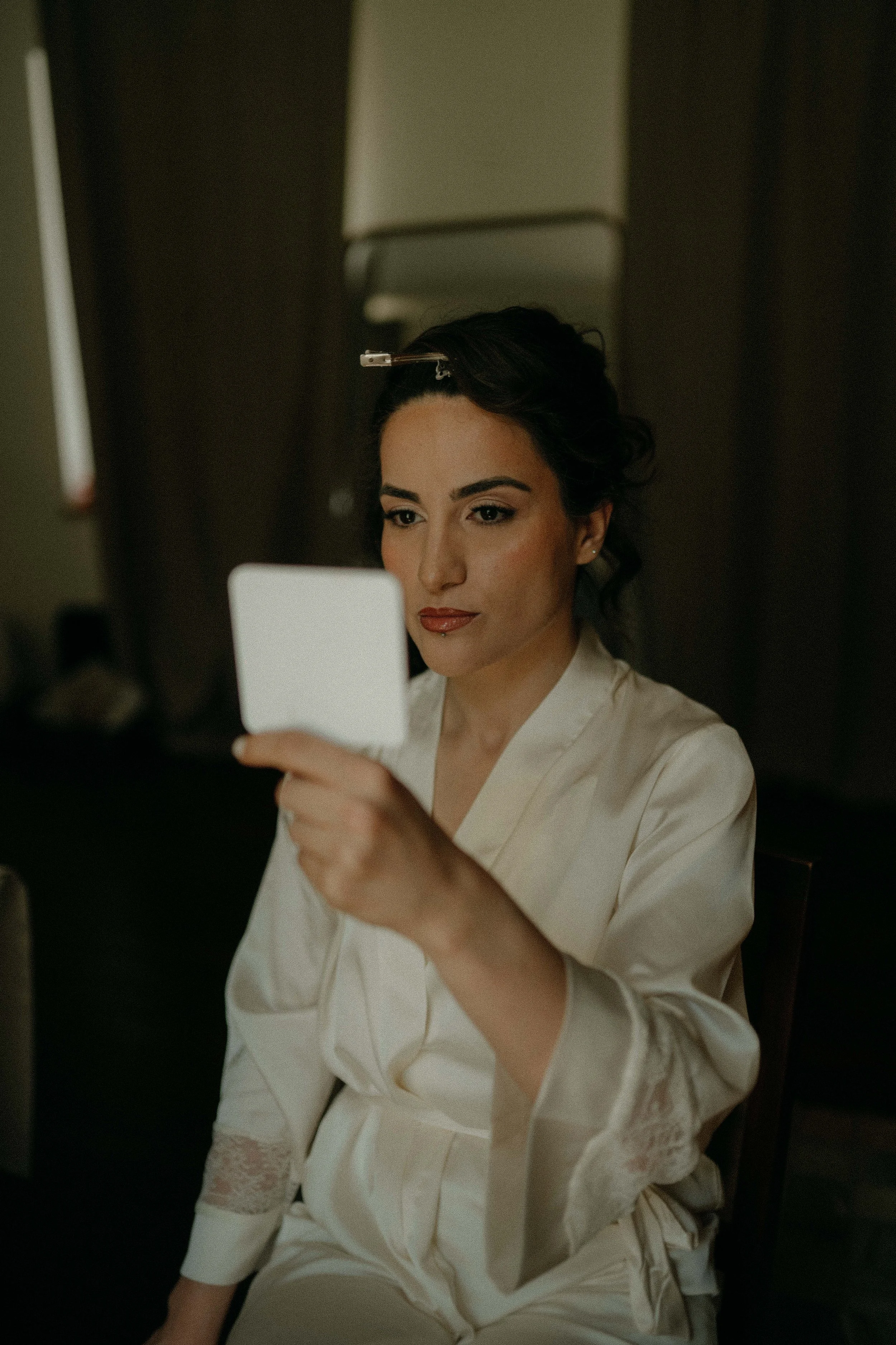 A woman sitting in a chair, dressed in white, getting her makeup done with a brush while looking into a mirror.