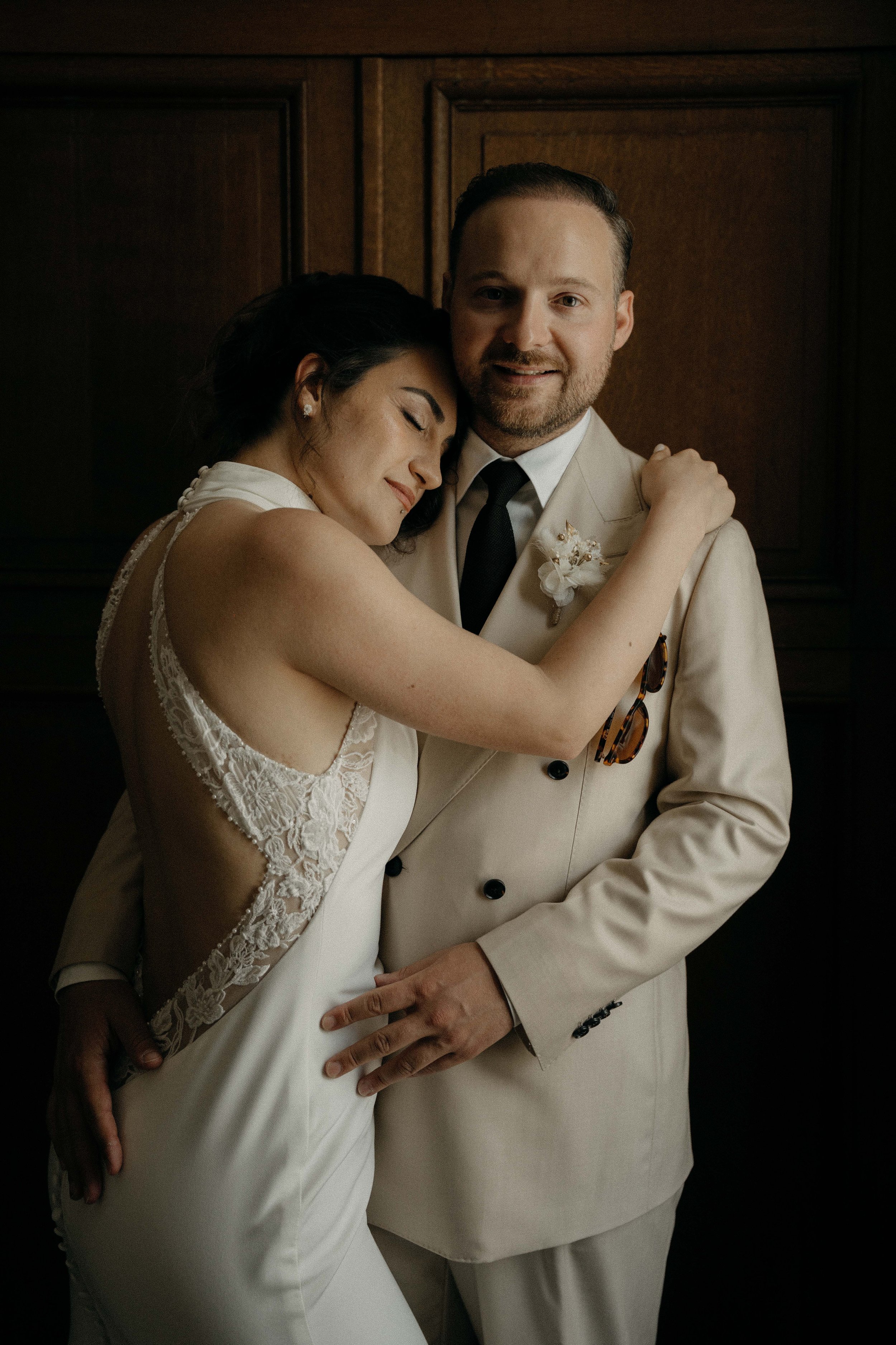 A bride and groom hugging each other, with the bride resting her head on the groom's shoulder. They are dressed in wedding attire, with the groom in a beige suit and the bride in a white lace gown. The background features dark wooden paneling.