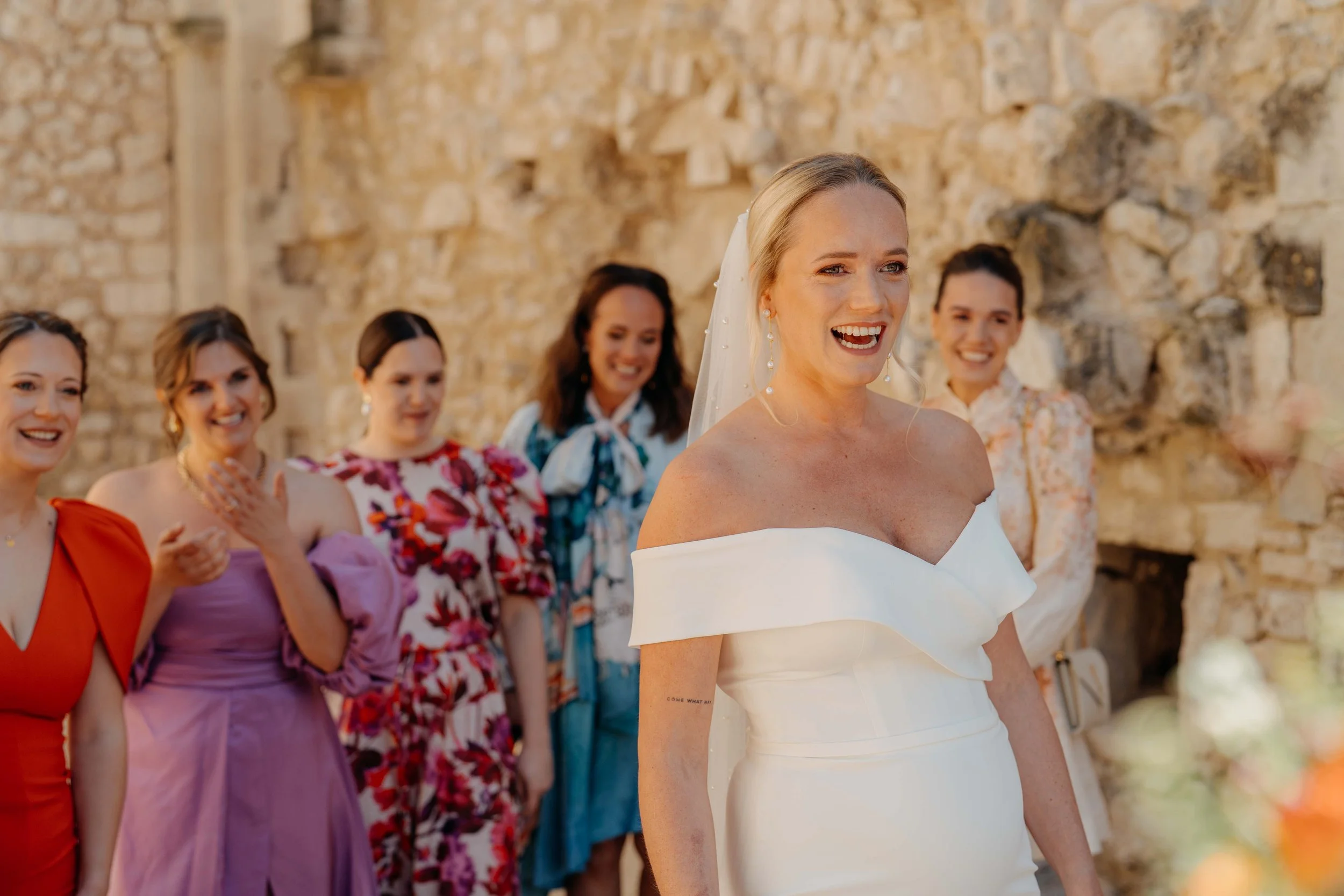 A bride in a white off-shoulder wedding dress smiling, surrounded by women in colorful dresses, at an outdoor wedding with a stone wall background.