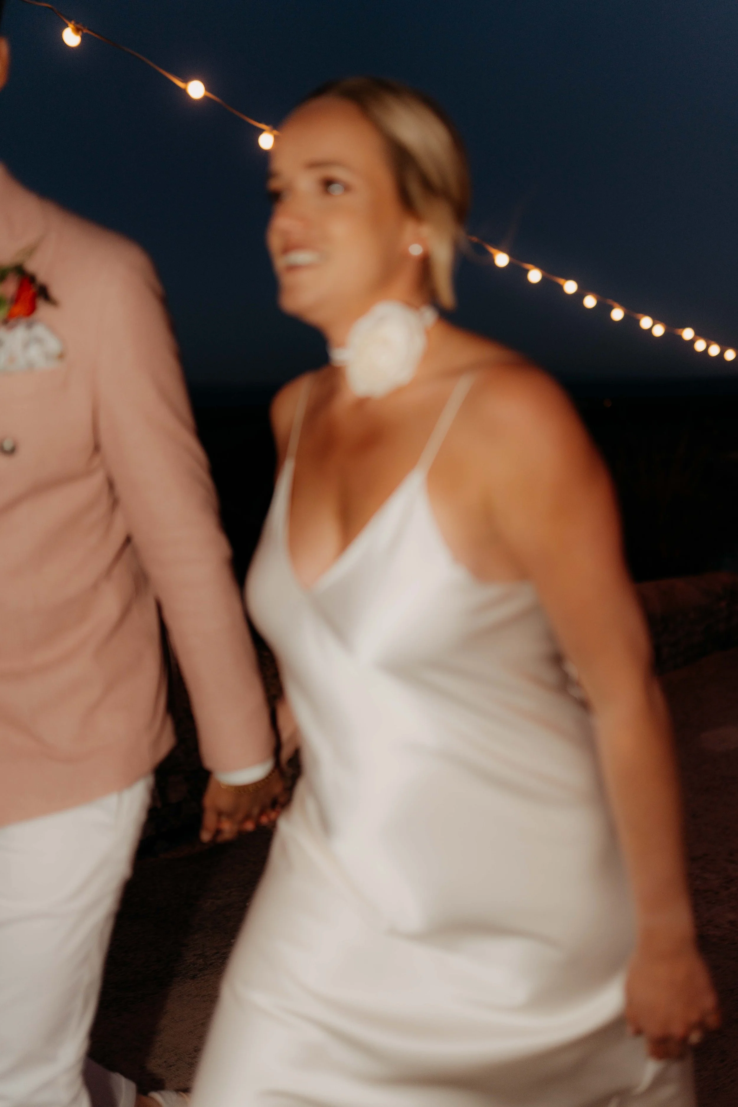 A woman smiling at a nighttime outdoor event with string lights in the background, wearing a white satin dress and a white floral choker necklace.