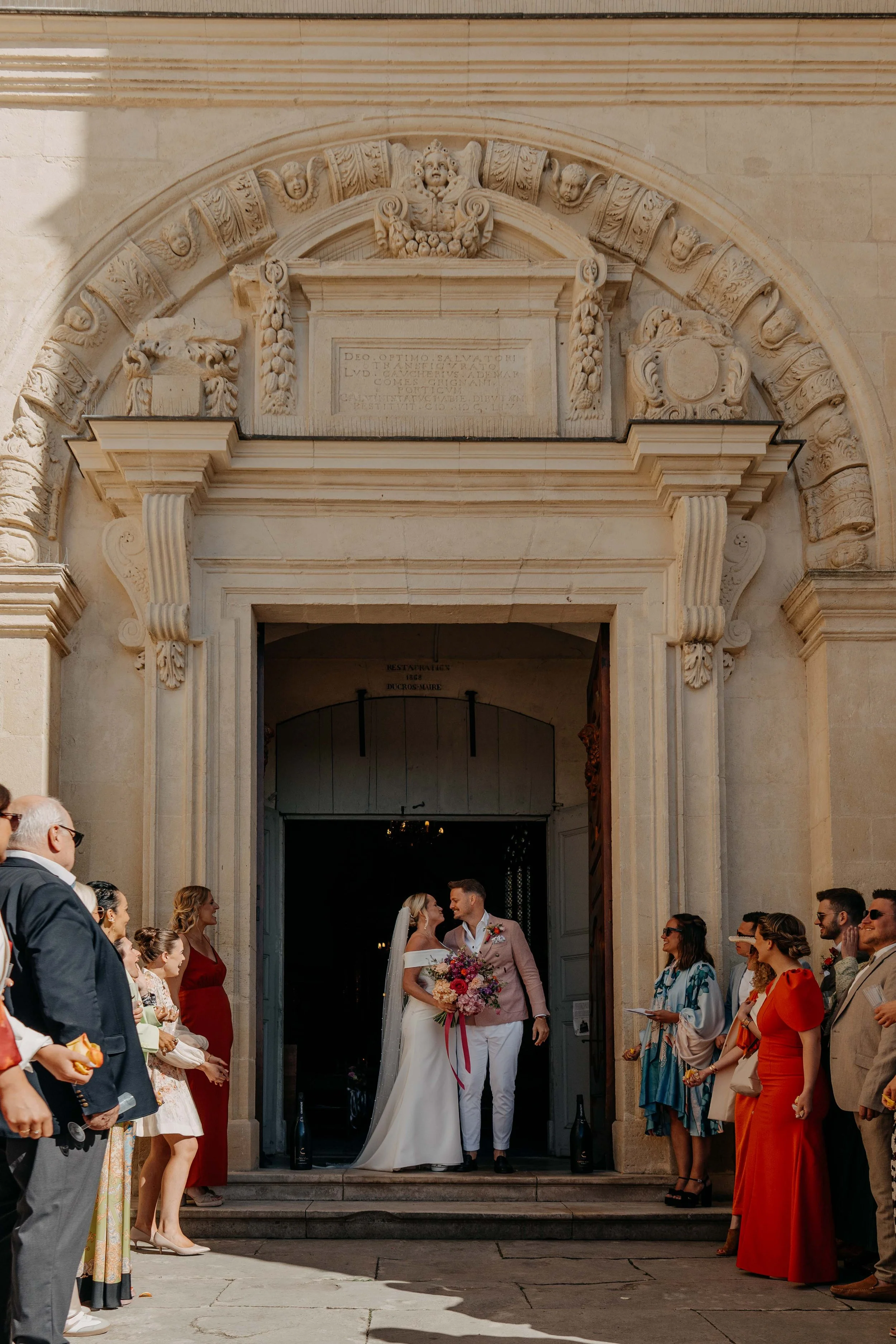 A bride and groom exiting a church with wedding guests lining the steps and standing nearby, with a large stone archway above entrance.