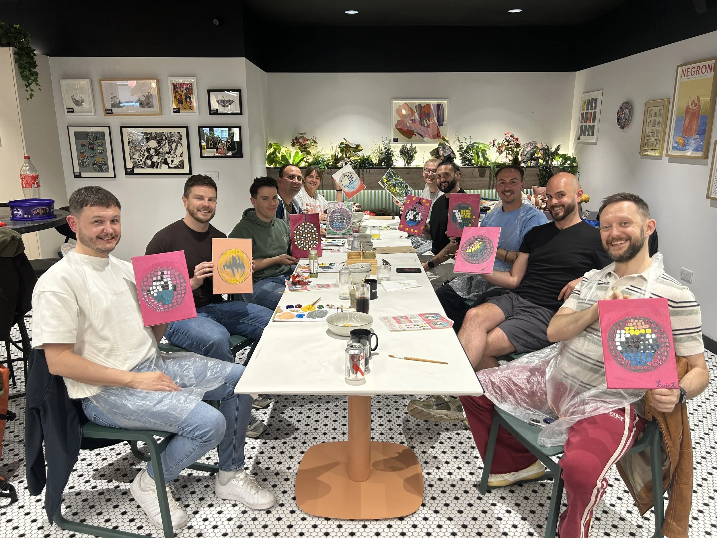 Group of people sitting around a table during a painting activity, holding up colorful paintings with circular patterns, in a room decorated with framed pictures and plants.