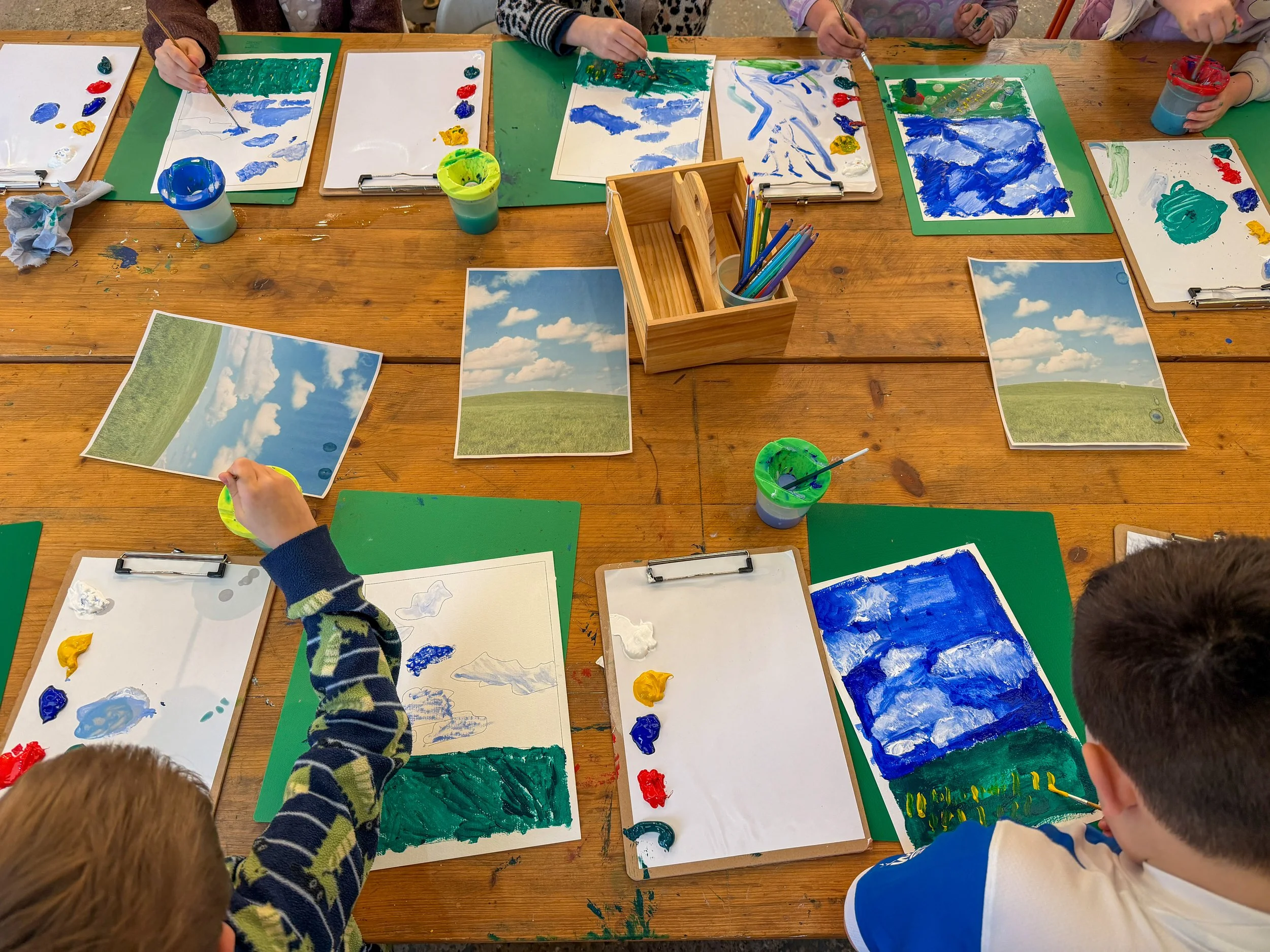 Children painting on white paper with various shades of blue, green, yellow, and red paint at a wooden table, with printed landscape pictures beside them.
