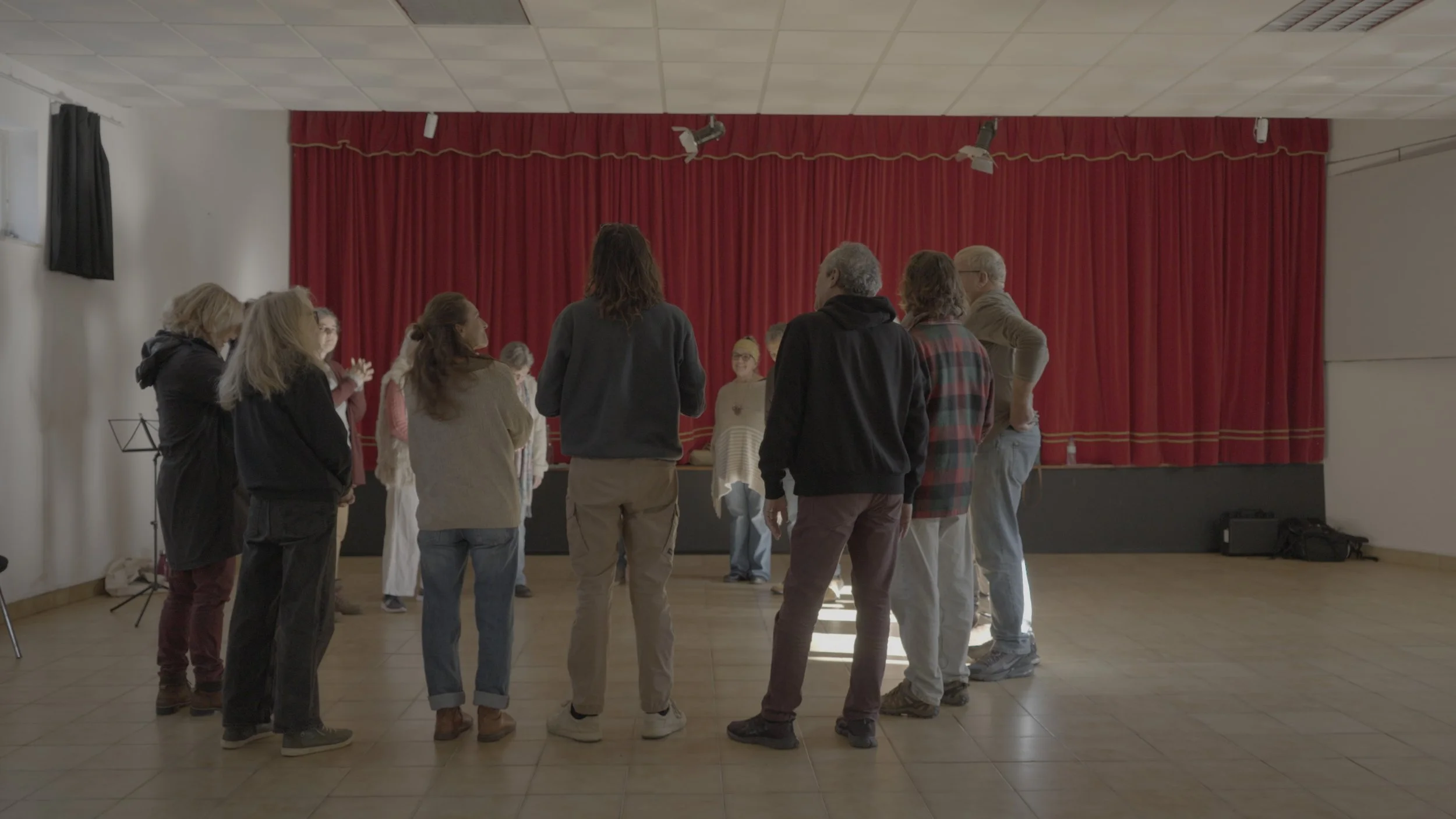 Grupo de várias pessoas ensaiando ou participando de atividade em um palco com cortina vermelha, em uma sala com teto de gesso e piso de azulejo.